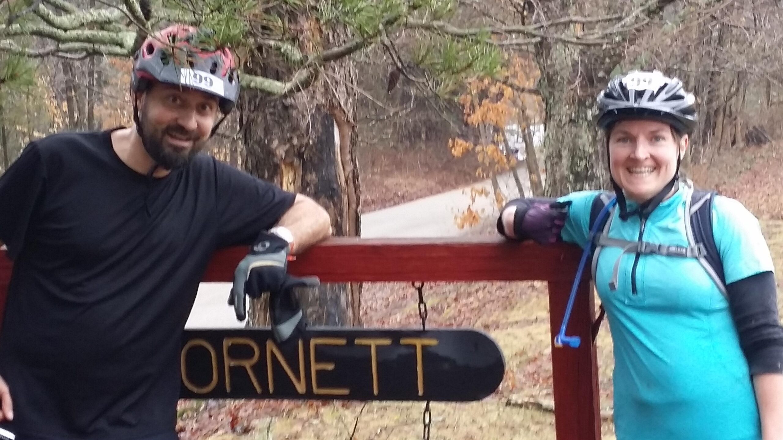 Two mountain bikers, a man and a woman, stand beside a wooden sign that reads "ORNETT." They are wearing helmets and biking gear, and both are smiling, enjoying a break during their ride. Trees and a road can be seen in the background, indicating a natural outdoor setting. Hickory Ridge mountain bike trail.
