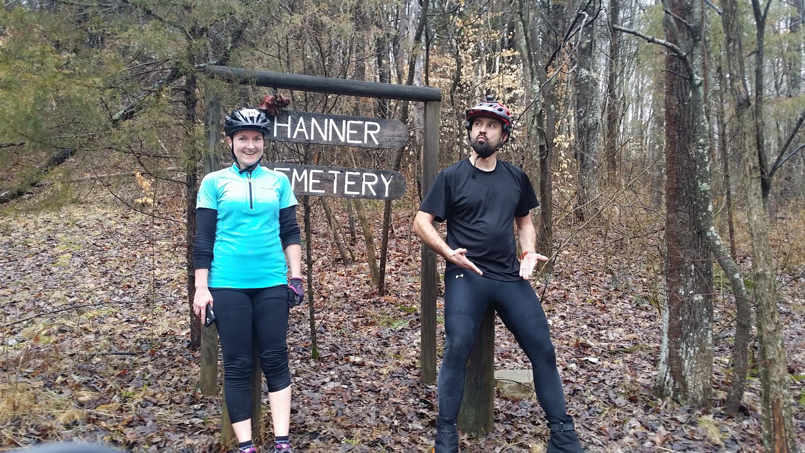 A man and a woman are standing in front of a wooden sign that reads "Hanner Cemetery." Both are wearing cycling helmets and outdoor athletic clothing. The background features a wooded area with trees and fallen leaves, suggesting a cool, possibly rainy day. The man is striking a playful pose, while the woman smiles at the camera. Hickory Ridge mountain bike trail.
