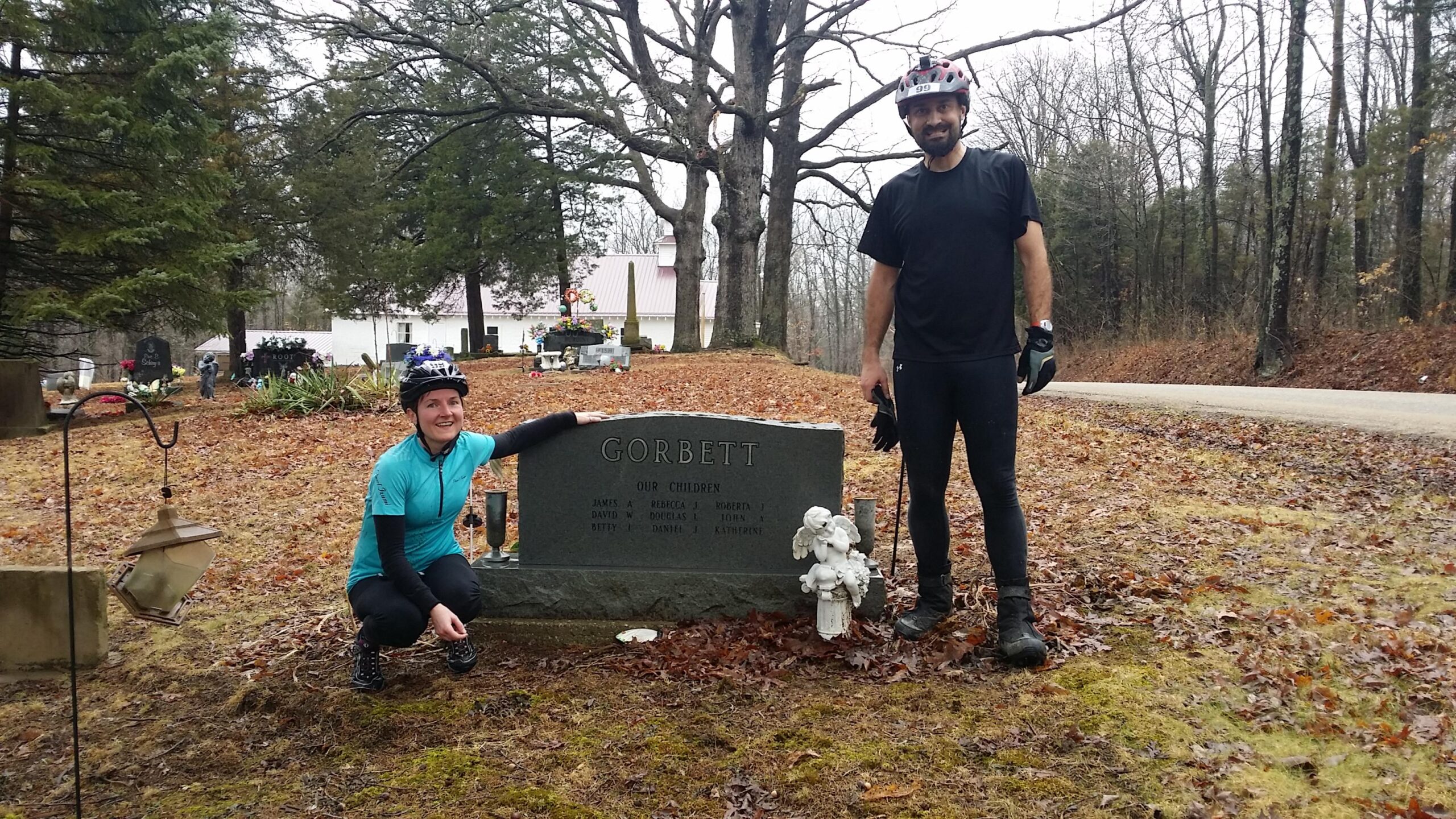 Two cyclists, a man and a woman, pose beside a gravestone marked "Gorbett," which lists family names. The woman is kneeling and smiling, wearing a turquoise cycling jersey and a helmet, while the man stands next to her in a black shirt and pants. The background includes a cemetery with scattered gravestones and trees, and the ground is covered with fallen leaves. Hickory Ridge mountain bike trail.
