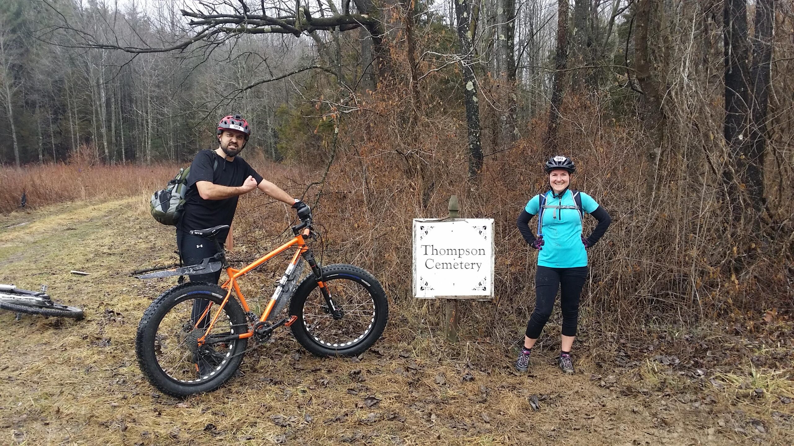 Alt text: Two mountain bikers posing beside a sign that reads "Thompson Cemetery." The man on the left is wearing a black shirt and helmet, flexing his arm, while the woman on the right is in a blue shirt, smiling with her hands on her hips. The background features trees and sparse brush. Hickory Ridge mountain bike trail.