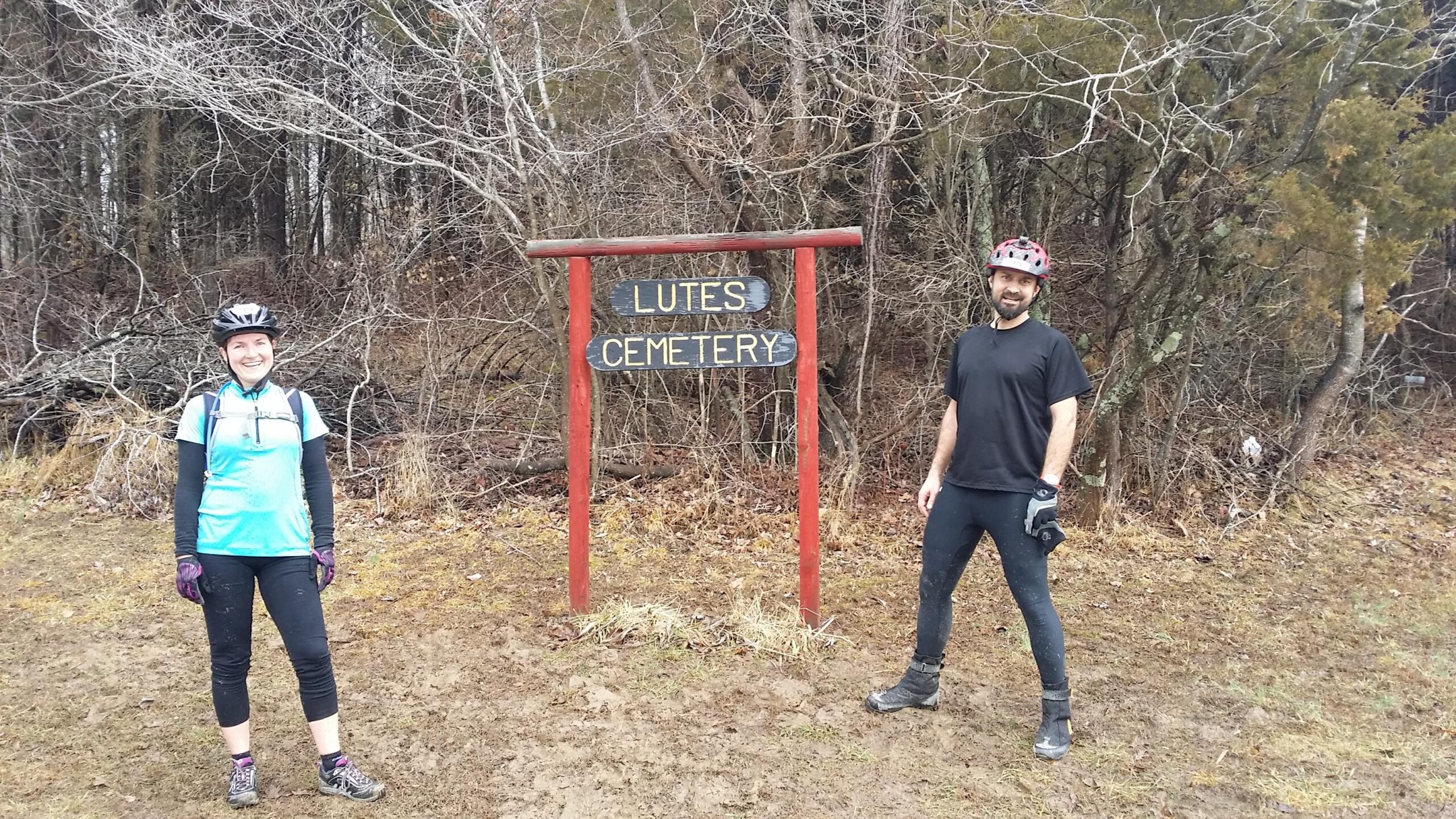 Two mountain bikers stand in front of a wooden sign that reads "Lutes Cemetery." The woman on the left is wearing a light blue cycling jersey, black leggings, and a helmet, smiling at the camera. The man on the right wears a black shirt and pants, with a red helmet and gloves, standing casually. The background features bare trees and a muddy area, suggesting a remote outdoor location. Hickory Ridge mountain bike trail.