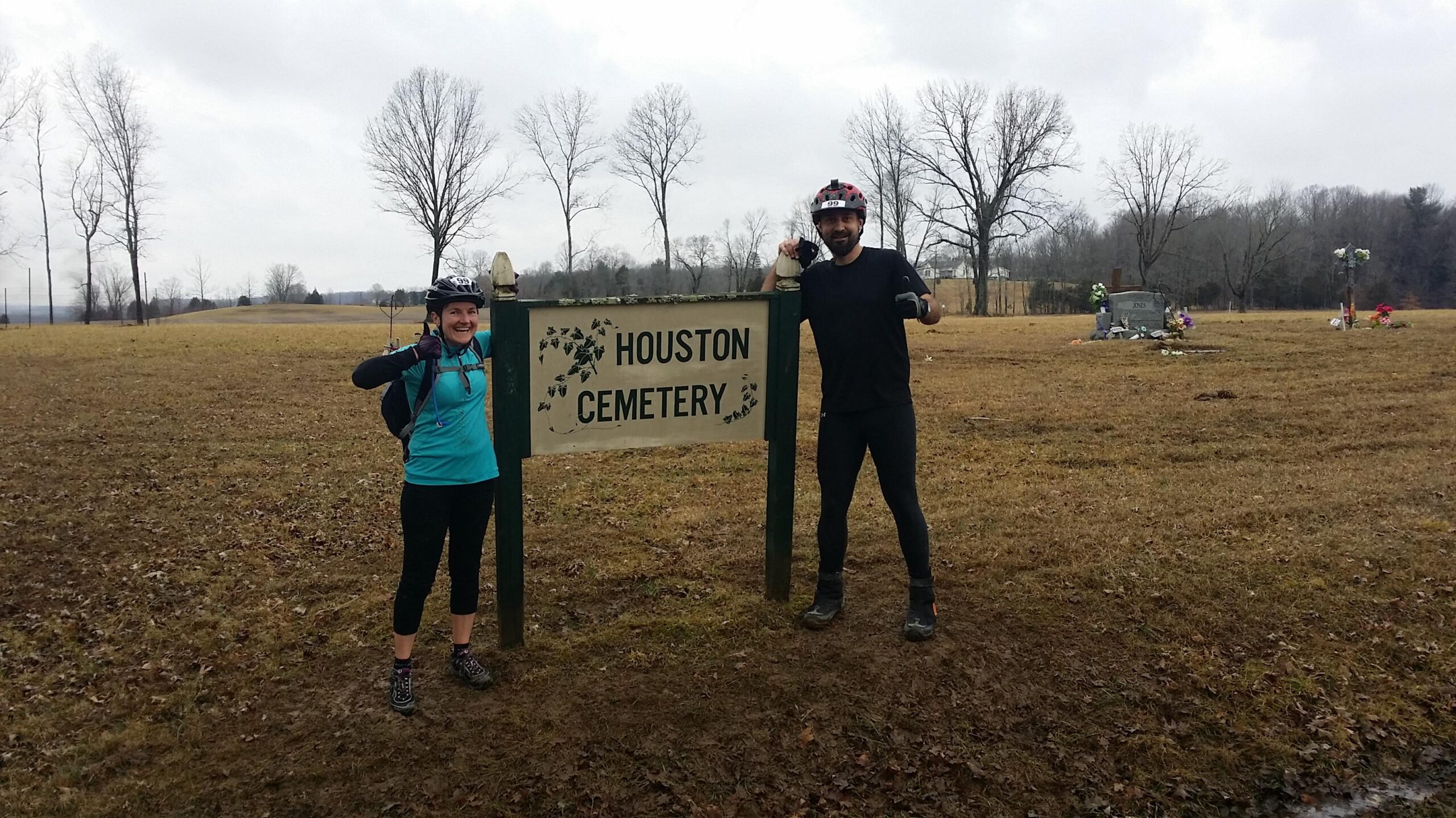 A man and a woman wearing biking gear stand beside a sign that reads "Houston Cemetery." The woman is giving a thumbs up, and the man is smiling next to her. The background features a grassy field with bare trees and another area with grave markers and flowers. The sky is overcast, indicating a cloudy day. Hickory Ridge mountain bike trail.