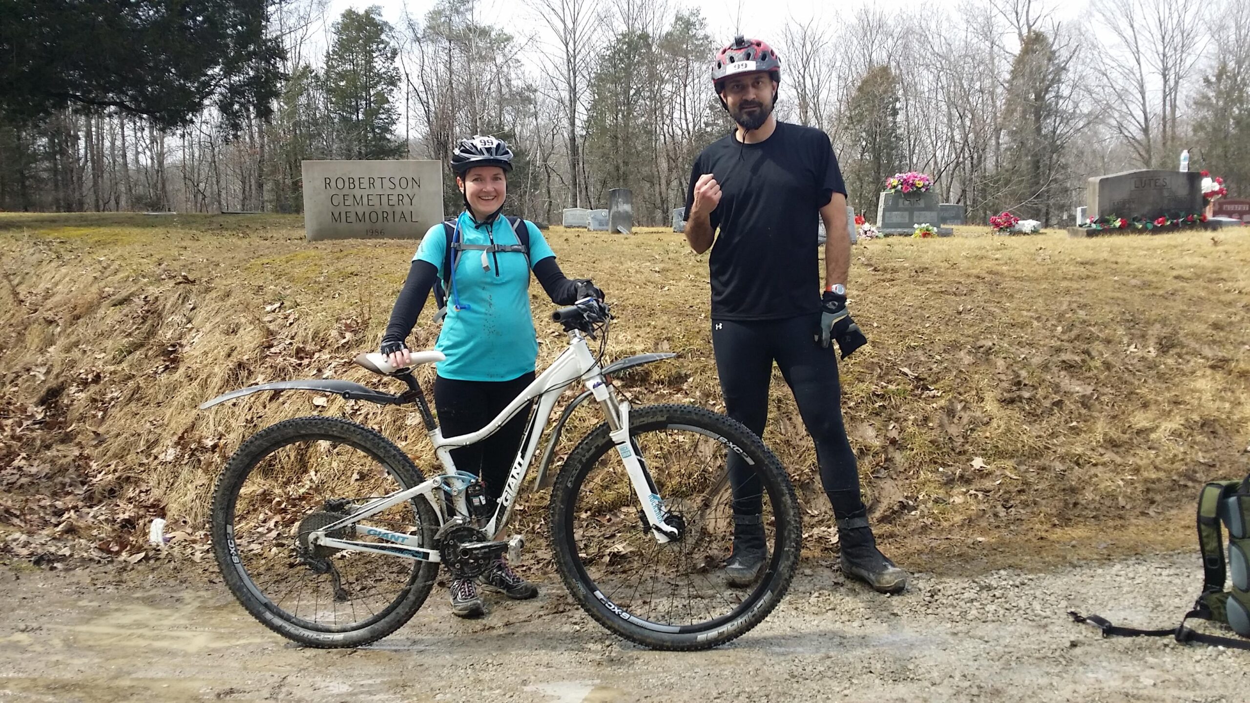 Two mountain bikers pose together beside a cemetery memorial. One rider, wearing a blue shirt and holding a bicycle, smiles at the camera. The other rider, dressed in black, raises a fist in a playful gesture. In the background, a stone sign reads "ROBERTSON CEMETERY MEMORIAL" surrounded by trees and headstones. The ground is muddy, indicating recent rain. Hickory Ridge mountain bike trail.
