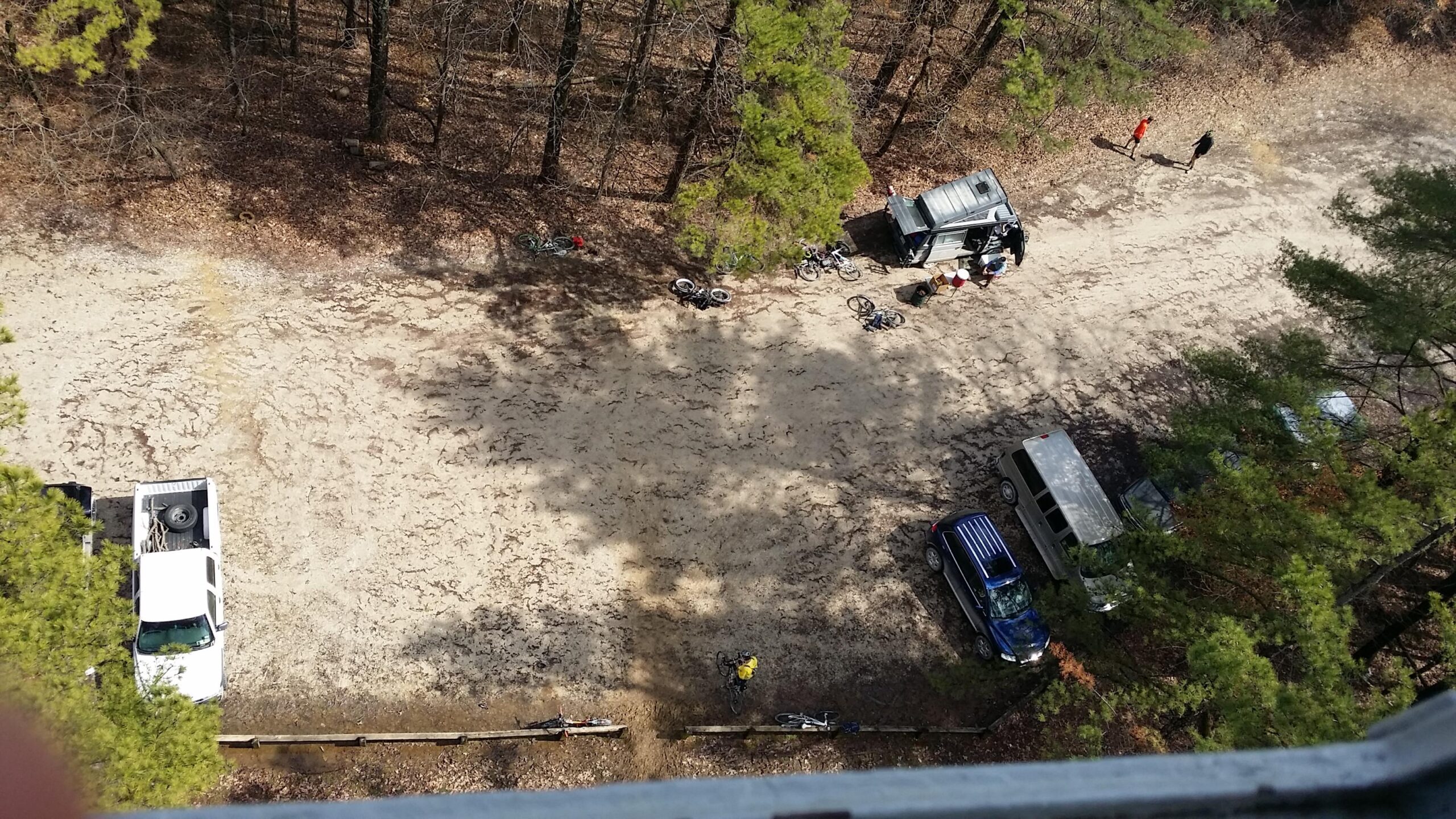 Aerial view of a forested area with a dirt road, showcasing several parked vehicles, bicycles scattered on the ground, and individuals walking. The ground is sandy and marked with tire tracks, surrounded by patches of dry leaves and trees. Hickory Ridge mountain bike trail.