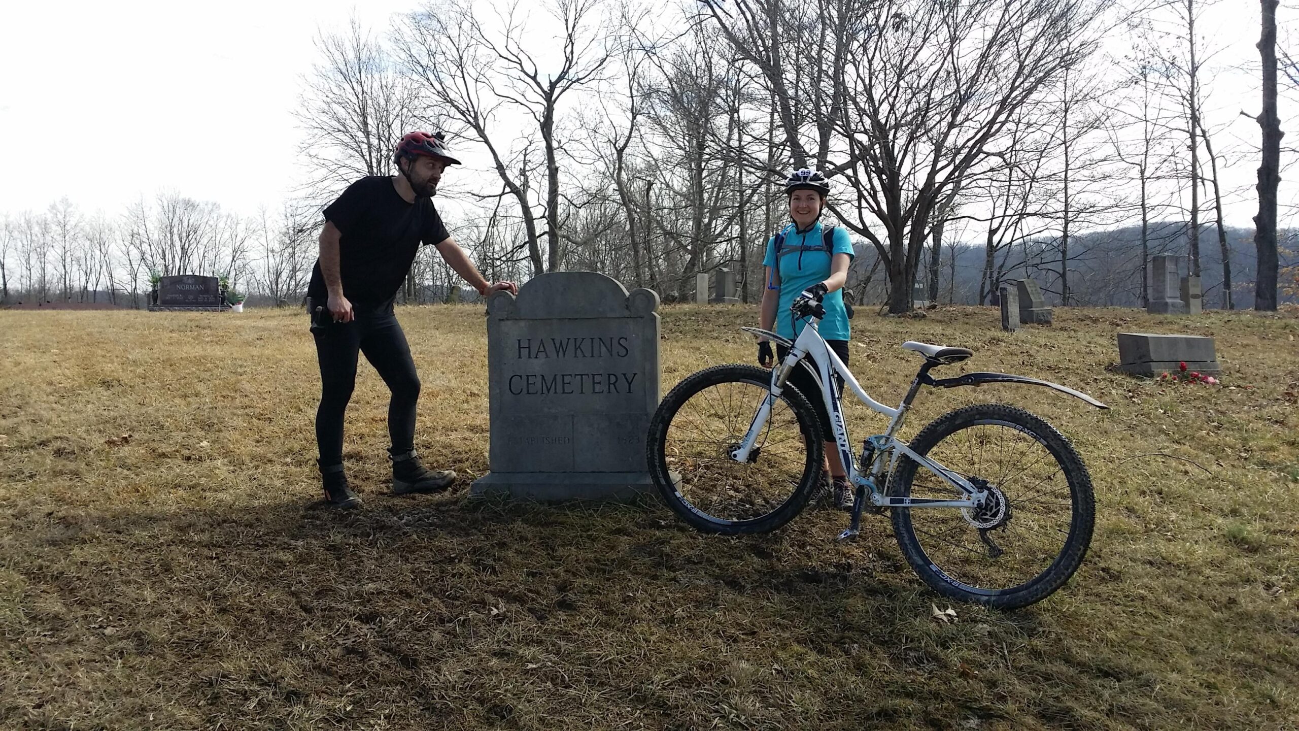 Two mountain bikers stand beside a gravestone marked "Hawkins Cemetery" in a grassy area surrounded by bare trees. One person is leaning on the gravestone while the other stands next to a bicycle. The scene captures the contrast of outdoor activity against a historical setting. Hickory Ridge mountain bike trail.