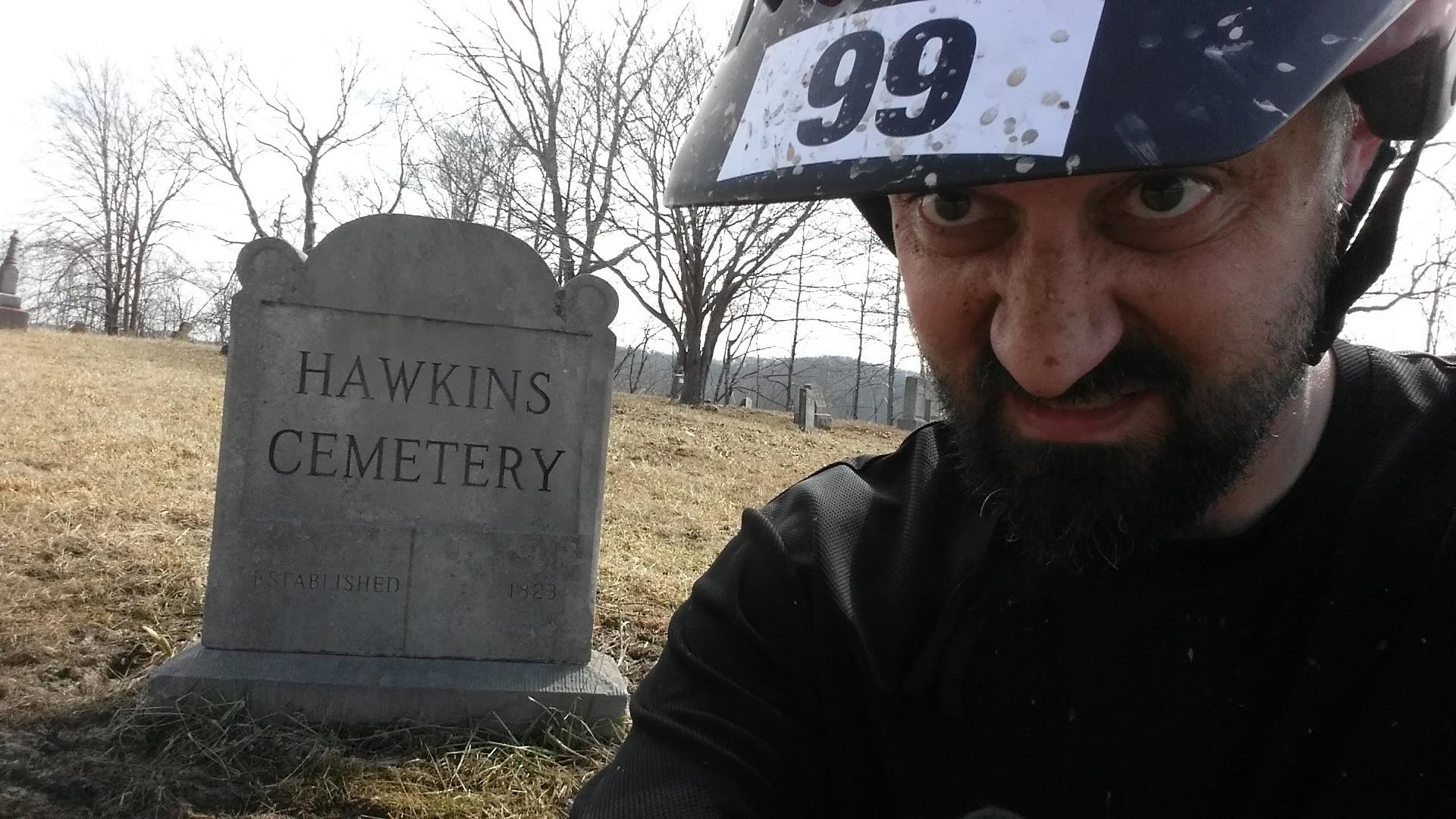 A person in a helmet with a race number takes a selfie in front of a gravestone that reads "HAWKINS CEMETERY," established in 1823. The background features bare trees and a grassy area typical of a cemetery setting. The individual's facial expression appears intense, adding an intriguing contrast to the peaceful cemetery scene. Hickory Ridge mountain bike trail.