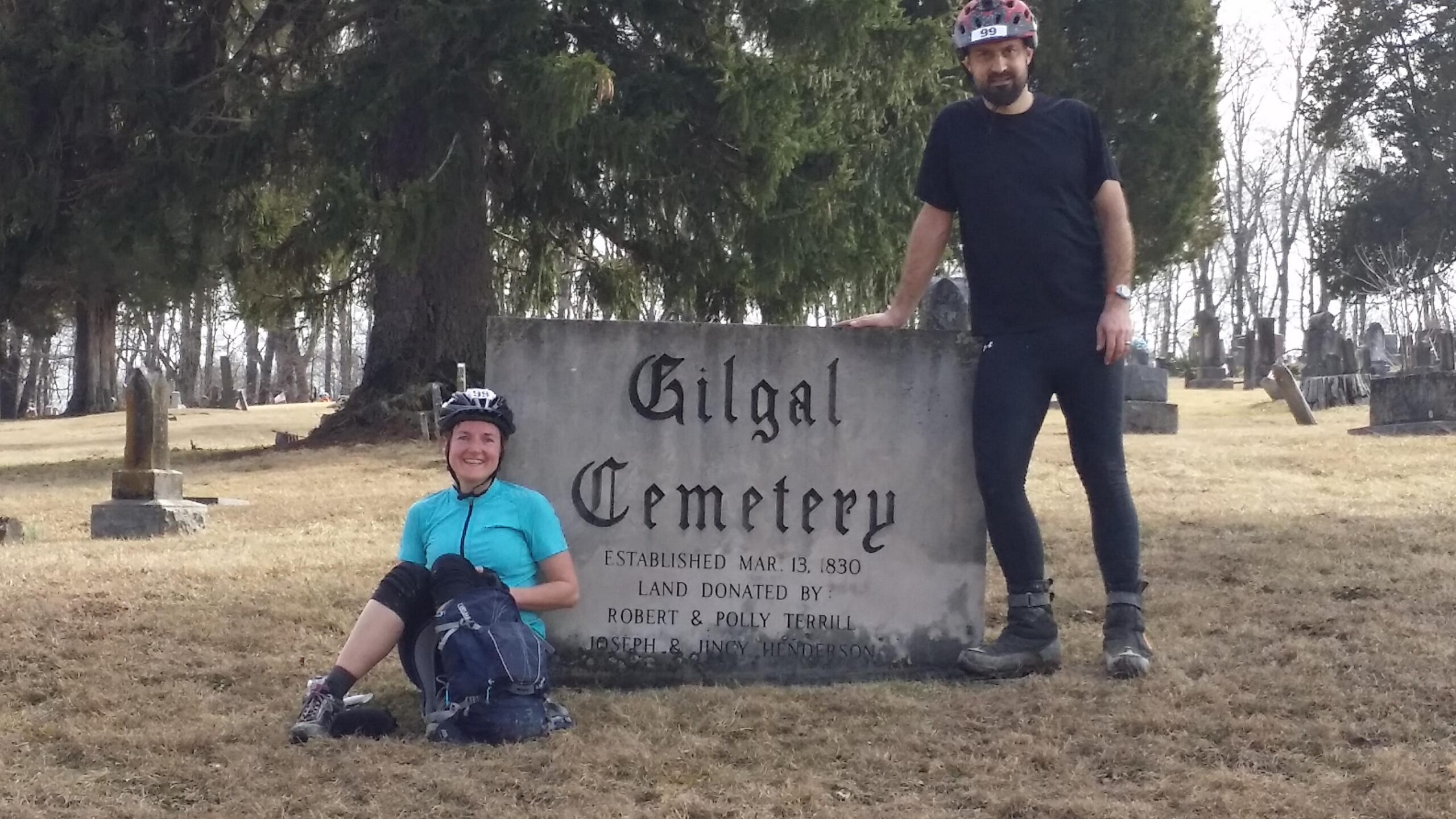 Two individuals, one sitting and one standing, pose in front of a sign marking "Gilgal Cemetery," which is established on March 13, 1830. The setting features grassy terrain and gravestones in the background, with trees surrounding the area. The person sitting wears a blue cycling shirt and helmet, while the standing person is dressed in black. Both appear to be enjoying their visit to the cemetery. Hickory Ridge mountain bike trail.