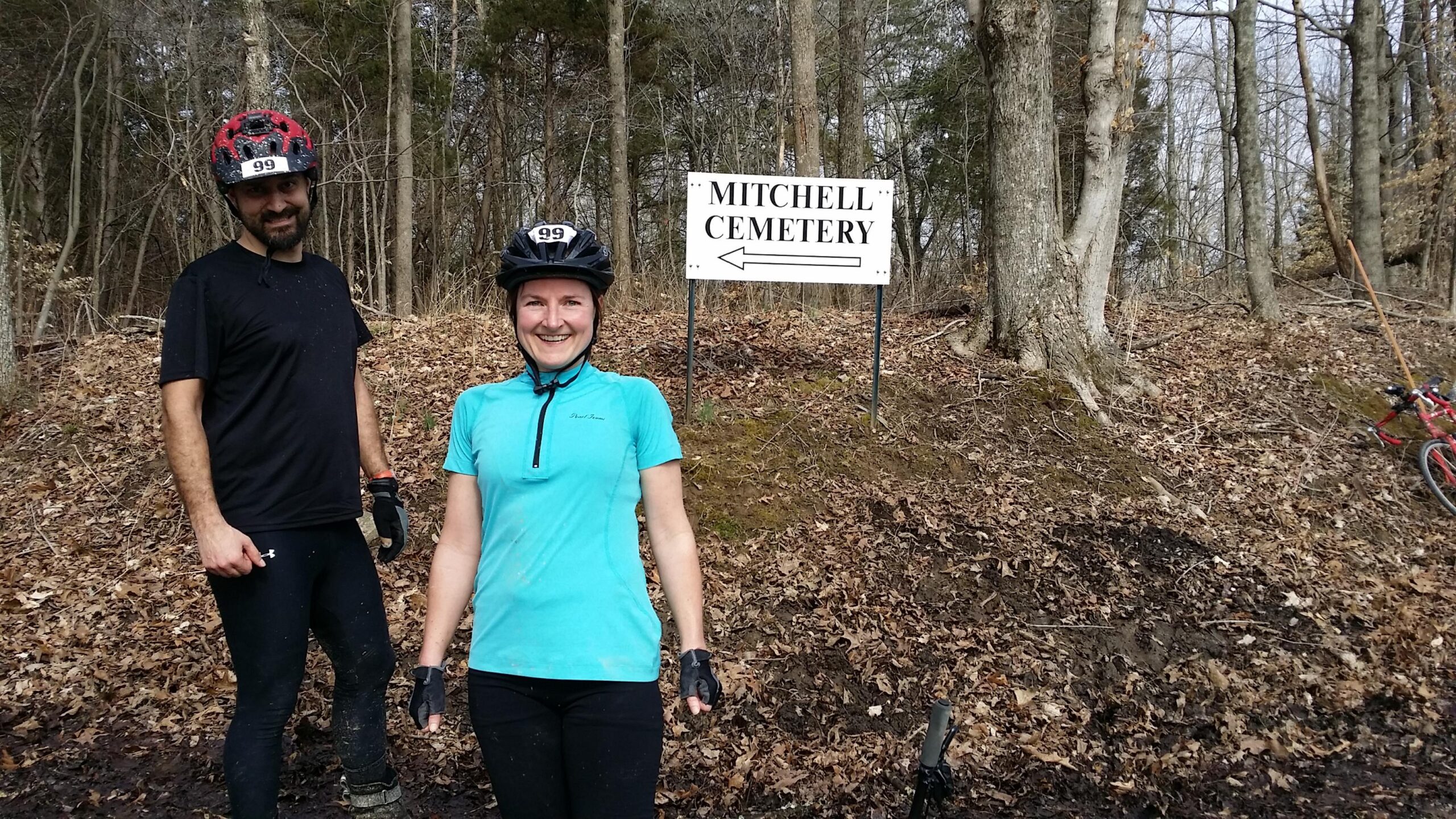 A man and a woman in cycling gear stand in a wooded area near a sign that reads "MITCHELL CEMETERY," with an arrow pointing to the left. They both wear helmets and are smiling, showcasing a sense of enjoyment outdoors. Fallen leaves cover the ground, and a bicycle is visible nearby. Hickory Ridge mountain bike trail.
