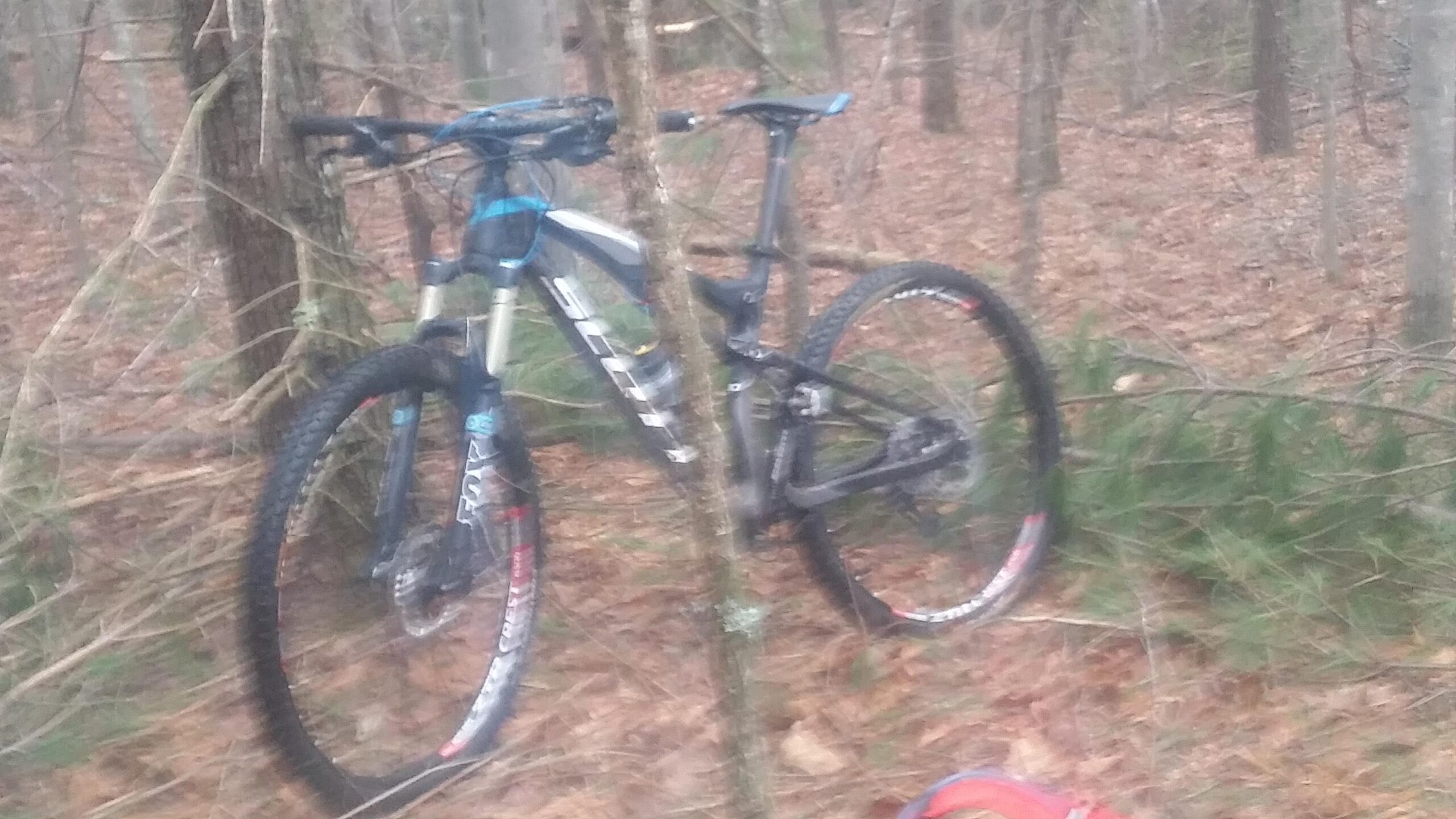 Two mountain bikes are leaning against trees in a forested area. The ground is covered with leaves and small branches, creating a natural setting. One bike has a blue and white color scheme, while the other is primarily black. A small backpack is partially visible in the foreground. Big South Fork mountain bike trail.