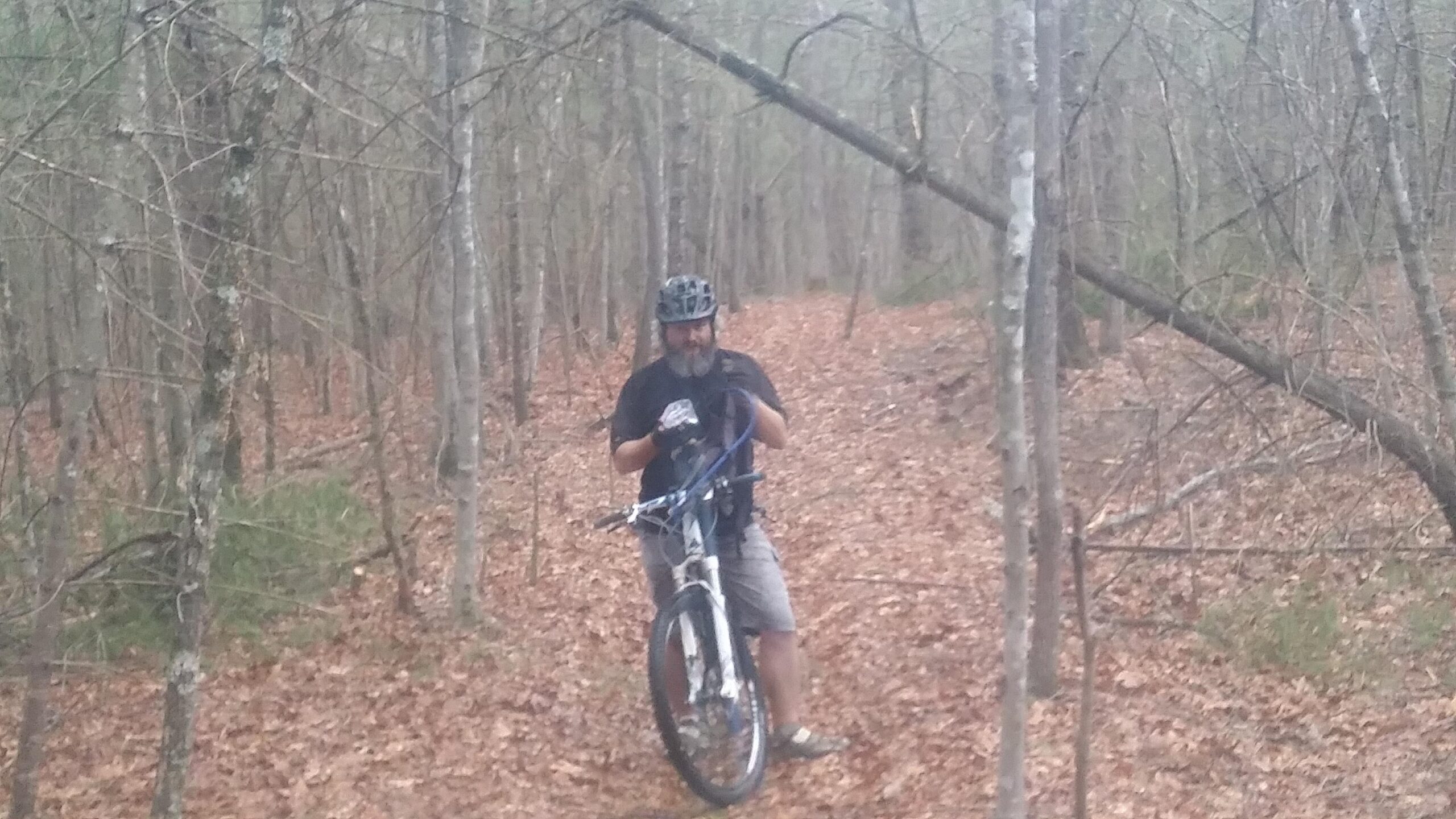A cyclist wearing a helmet and gloves stands on a dirt trail in a forest, holding a mountain bike. The ground is covered with fallen leaves, and trees with bare branches surround the area, creating a natural, wooded setting. Big South Fork mountain bike trail.