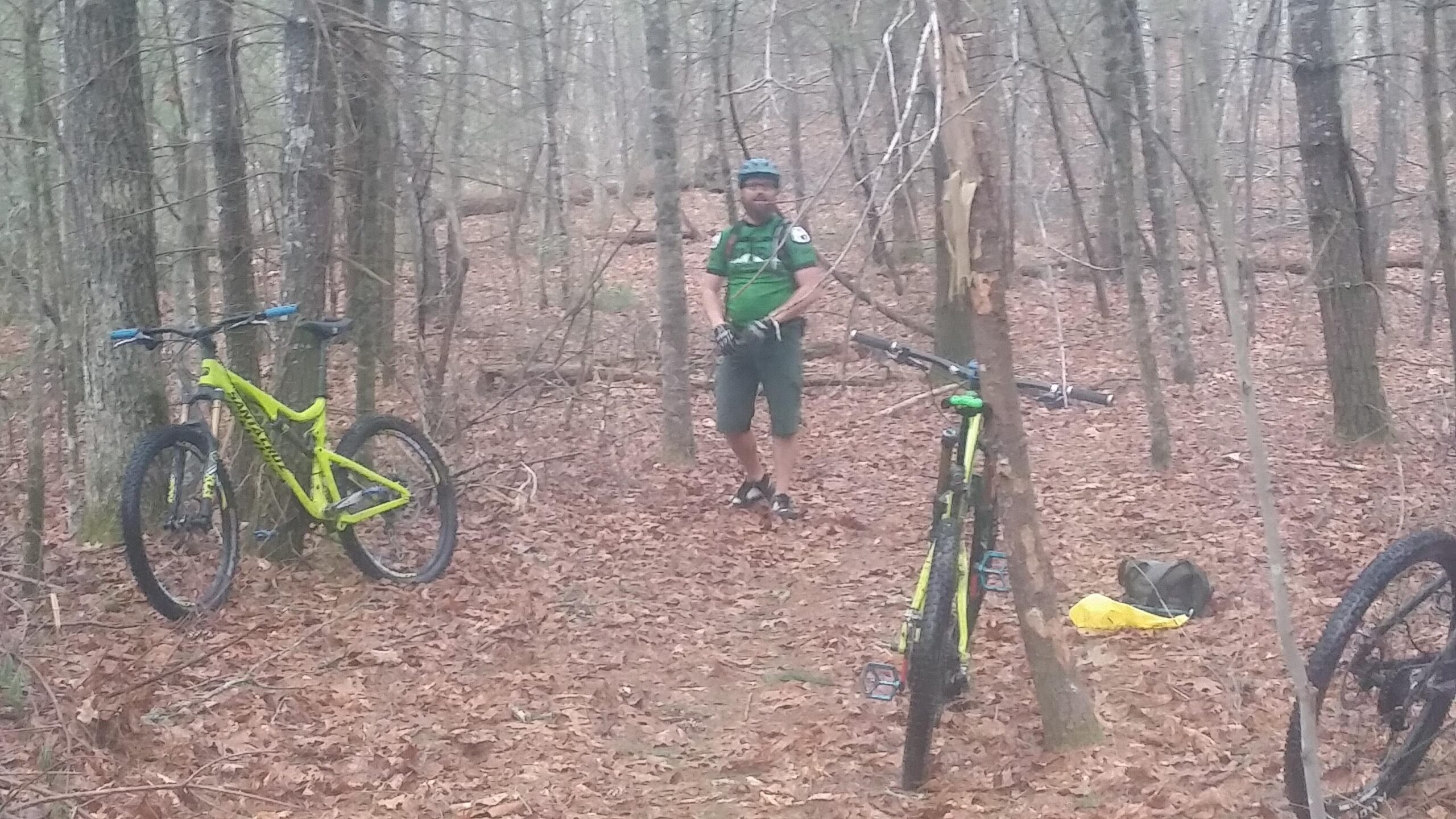 A man wearing a green shirt and helmet stands in a wooded area, surrounded by tall trees and fallen leaves. Two bright yellow and green mountain bikes are propped against trees nearby, and a backpack can be seen on the ground. The scene captures a moment of rest or pause while biking in nature. Big South Fork mountain bike trail.