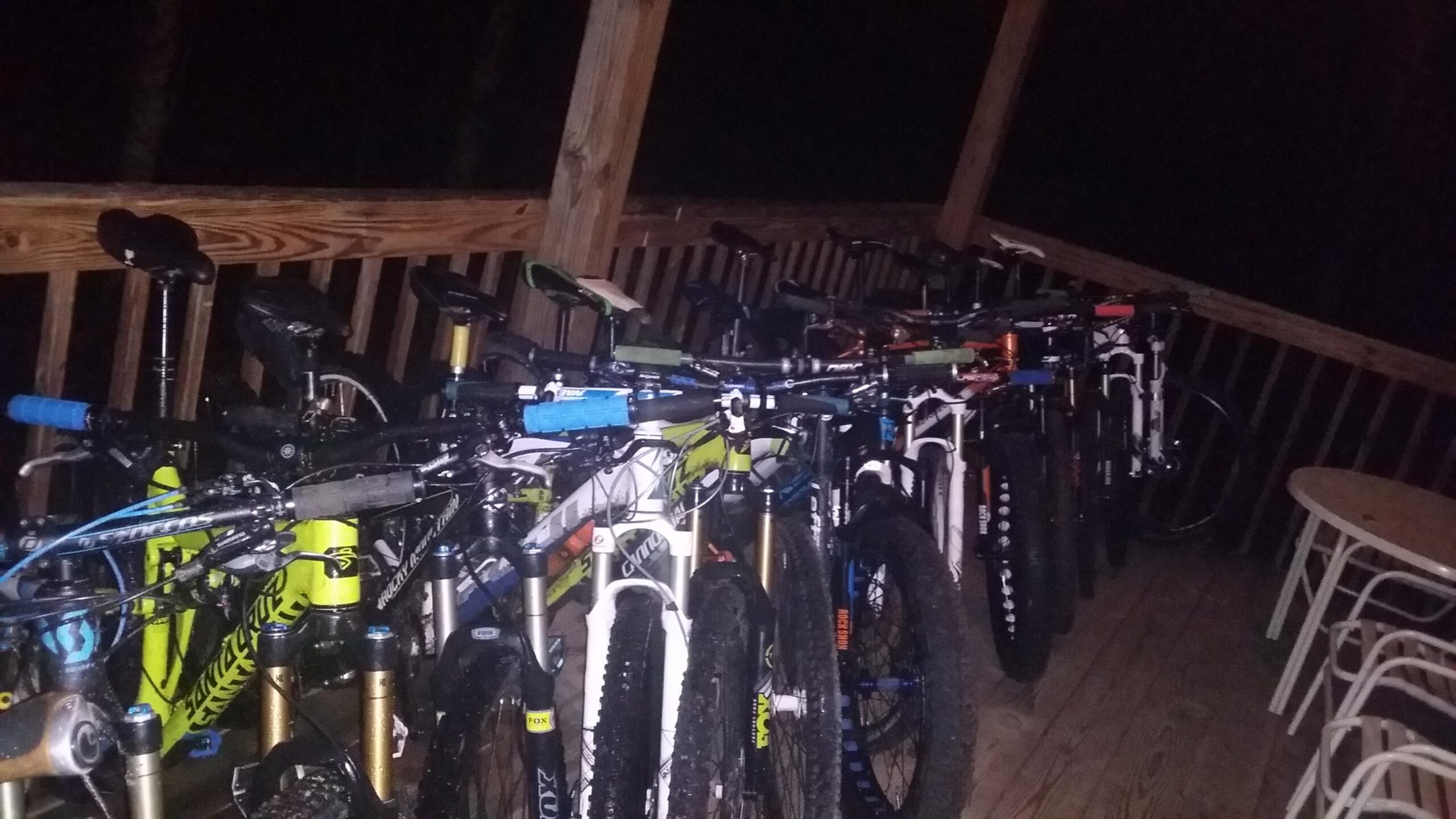 A collection of bicycles is parked on a wooden deck at night, with various colors and designs visible. Some bikes have brightly colored handlebars, while others appear to have distinct features like suspension forks and large tires. In the foreground, a white bike stands out, and a small table and chairs can be seen in the lower part of the image. The area is dimly lit, suggesting an outdoor setting. Big South Fork mountain bike trail.