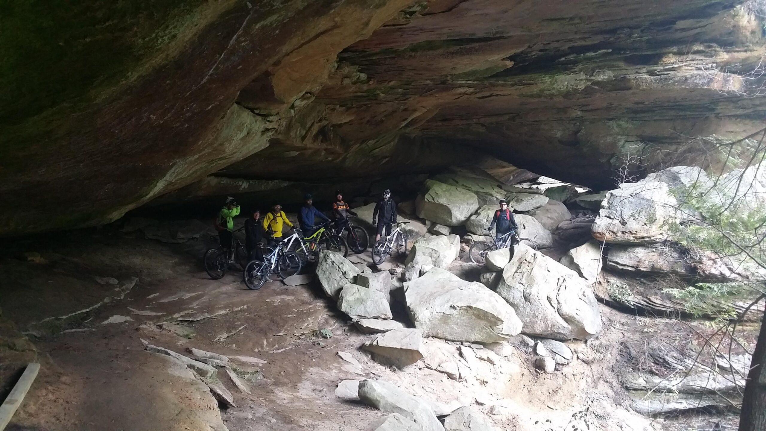 A group of seven mountain bikers stands among large rocks beneath a natural rock overhang in a wooded area. The cyclists are wearing various biking gear and helmets, and their bicycles are positioned beside them. The scene features rugged terrain with a mix of dirt and stone, and greenery is visible in the background. Big South Fork mountain bike trail.