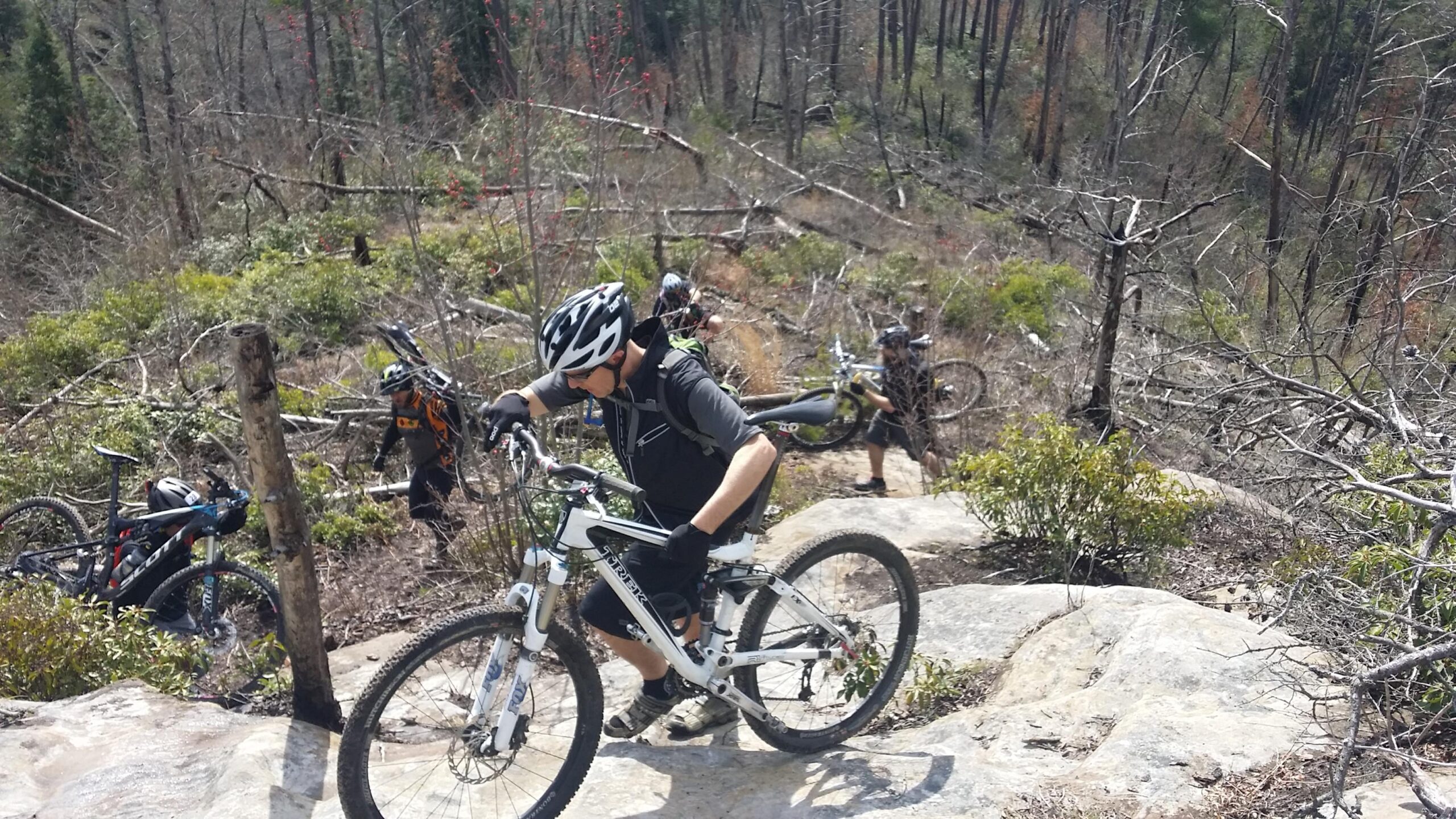 A group of mountain bikers navigating a rocky trail in a forested area. One cyclist is pushing their bike up a steep section of the path, while two others can be seen in the background. The scene features a mix of greenery and fallen trees, indicating a rugged environment. Big South Fork mountain bike trail.