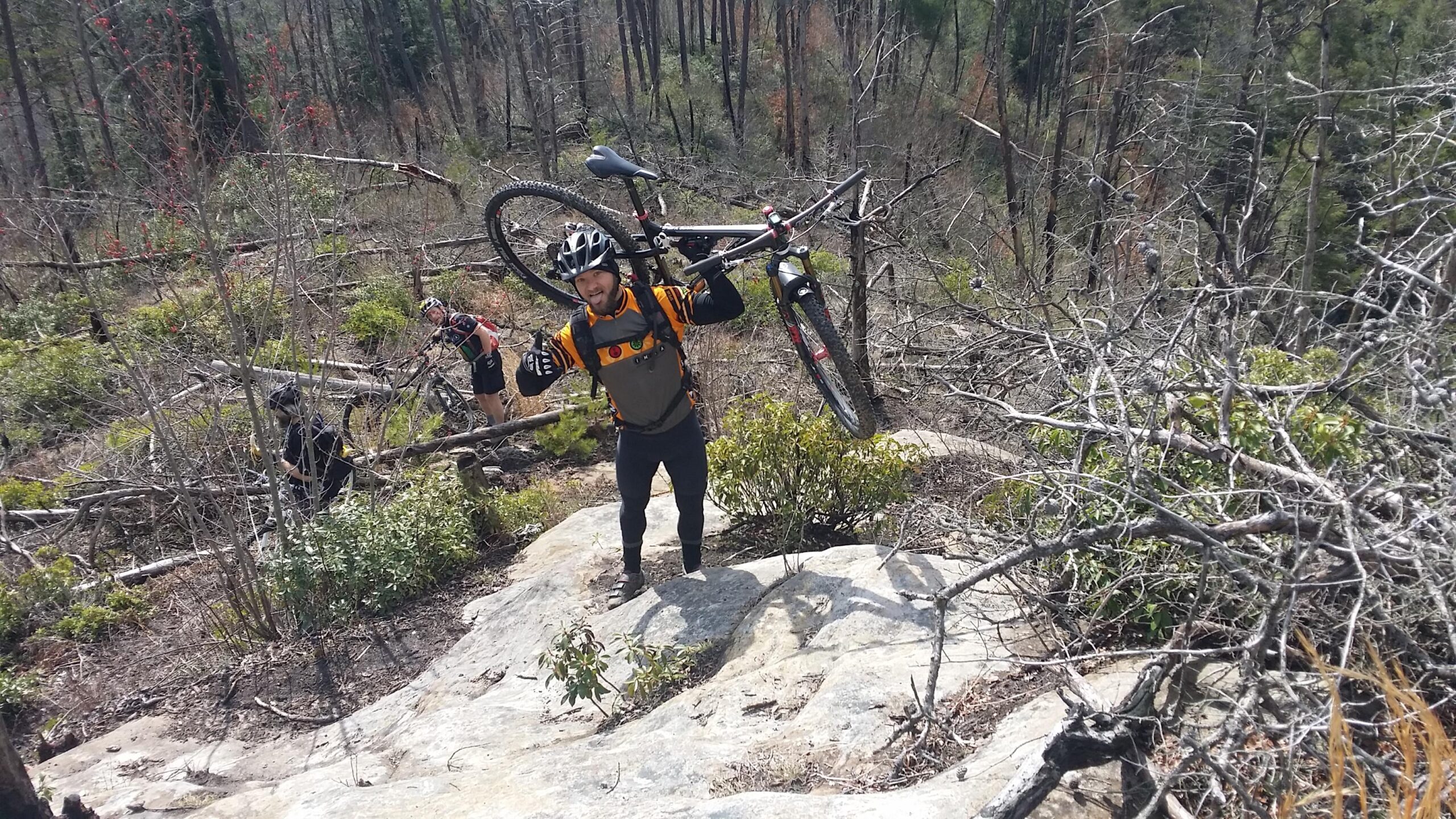 A mountain biker wearing a helmet and sporting gear is carrying a bicycle over rough terrain. He stands on a rocky surface amidst a landscape with scattered branches and shrubs. In the background, another person can be seen navigating the trail. The scene suggests an outdoor adventure in a natural setting. Big South Fork mountain bike trail.