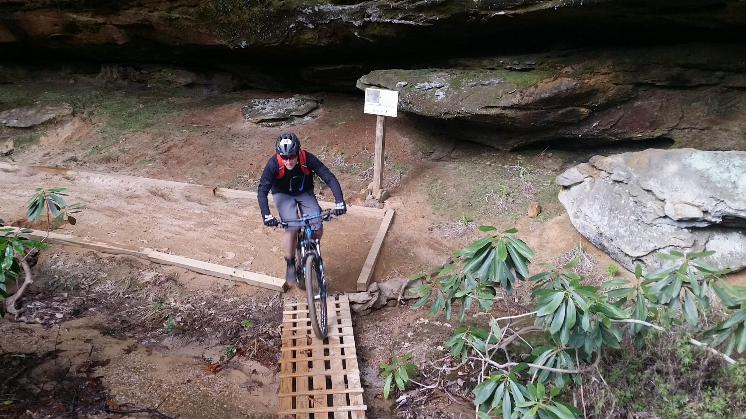 A mountain biker riding over a wooden bridge on a dirt trail in a forested area, with rocky outcrops and a sign visible in the background. Lush green plants and underbrush surround the trail, indicating a natural outdoor setting. Big South Fork mountain bike trail.