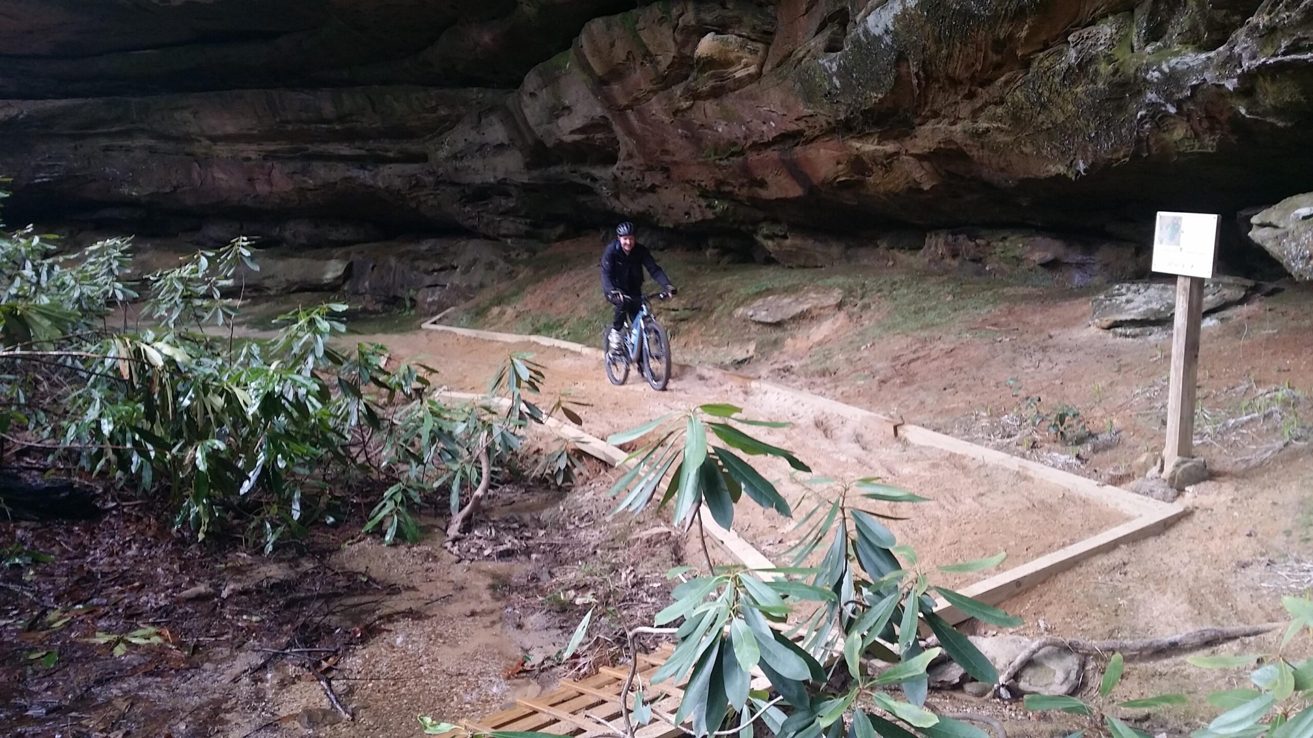 A mountain biker riding along a dirt trail beneath a rocky overhang, surrounded by greenery and small shrubs. A wooden sign is visible in the background, indicating the trail route. Big South Fork mountain bike trail.
