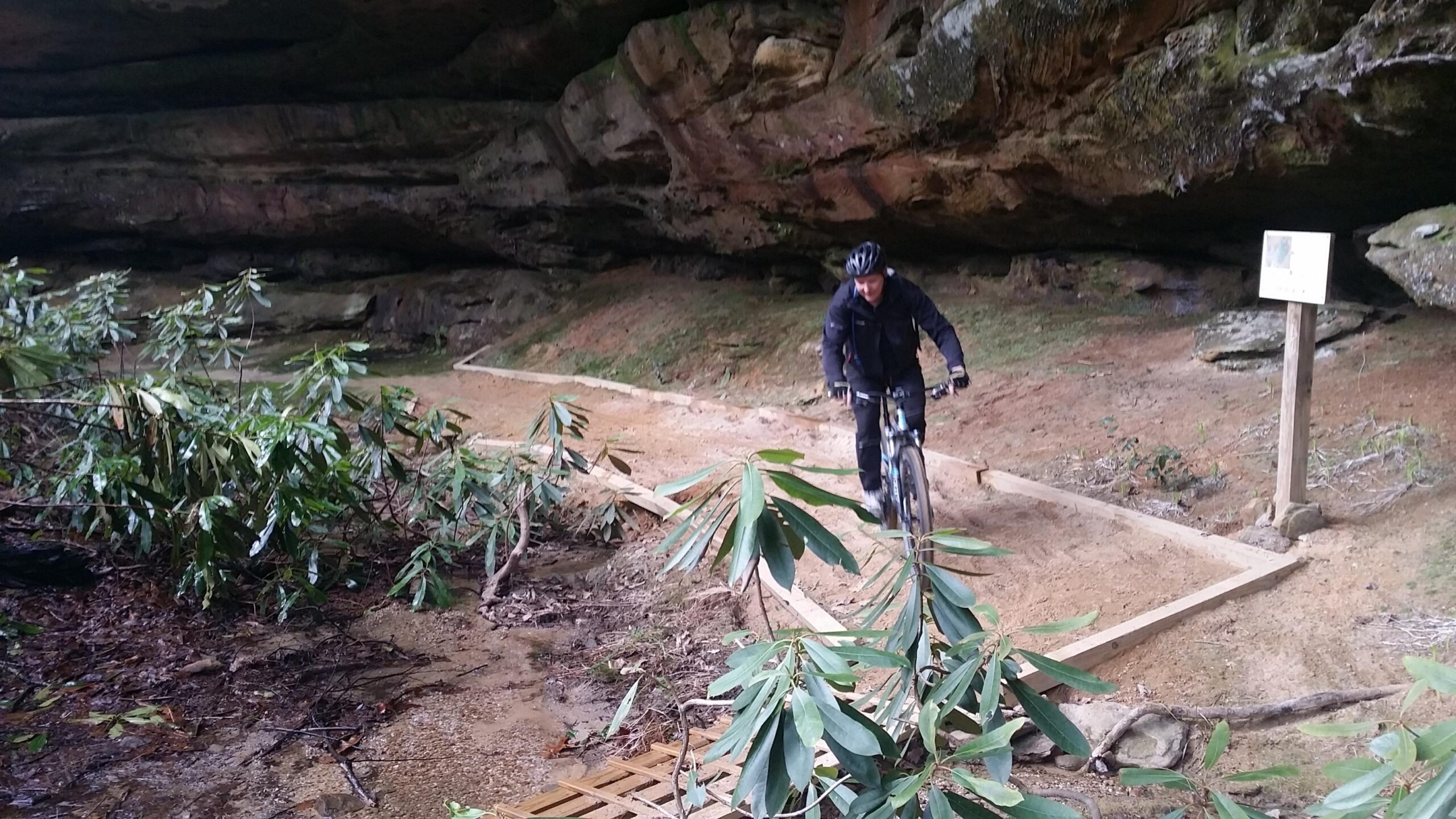 A mountain biker rides on a sandy trail beneath a rocky overhang, surrounded by green foliage. A wooden path curves along the trail, and a signpost is visible in the background. The scene depicts an outdoor recreational area. Big South Fork mountain bike trail.