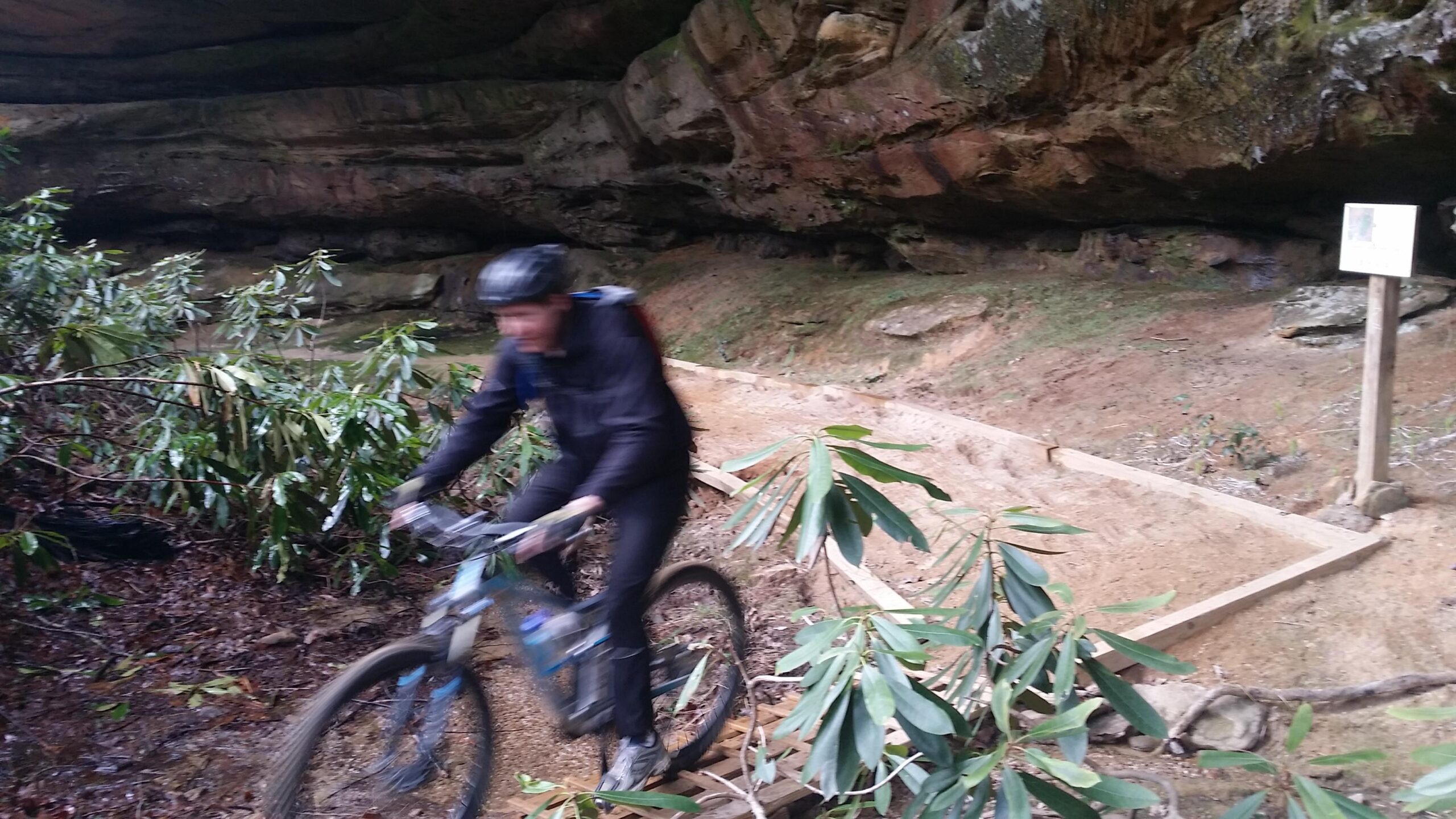 A mountain biker rides swiftly along a dirt path under a rocky overhang, surrounded by green foliage. The trail is narrow and features wooden edging, while a sign is visible in the background. The scene captures the excitement of biking in a natural, forested environment. Big South Fork mountain bike trail.