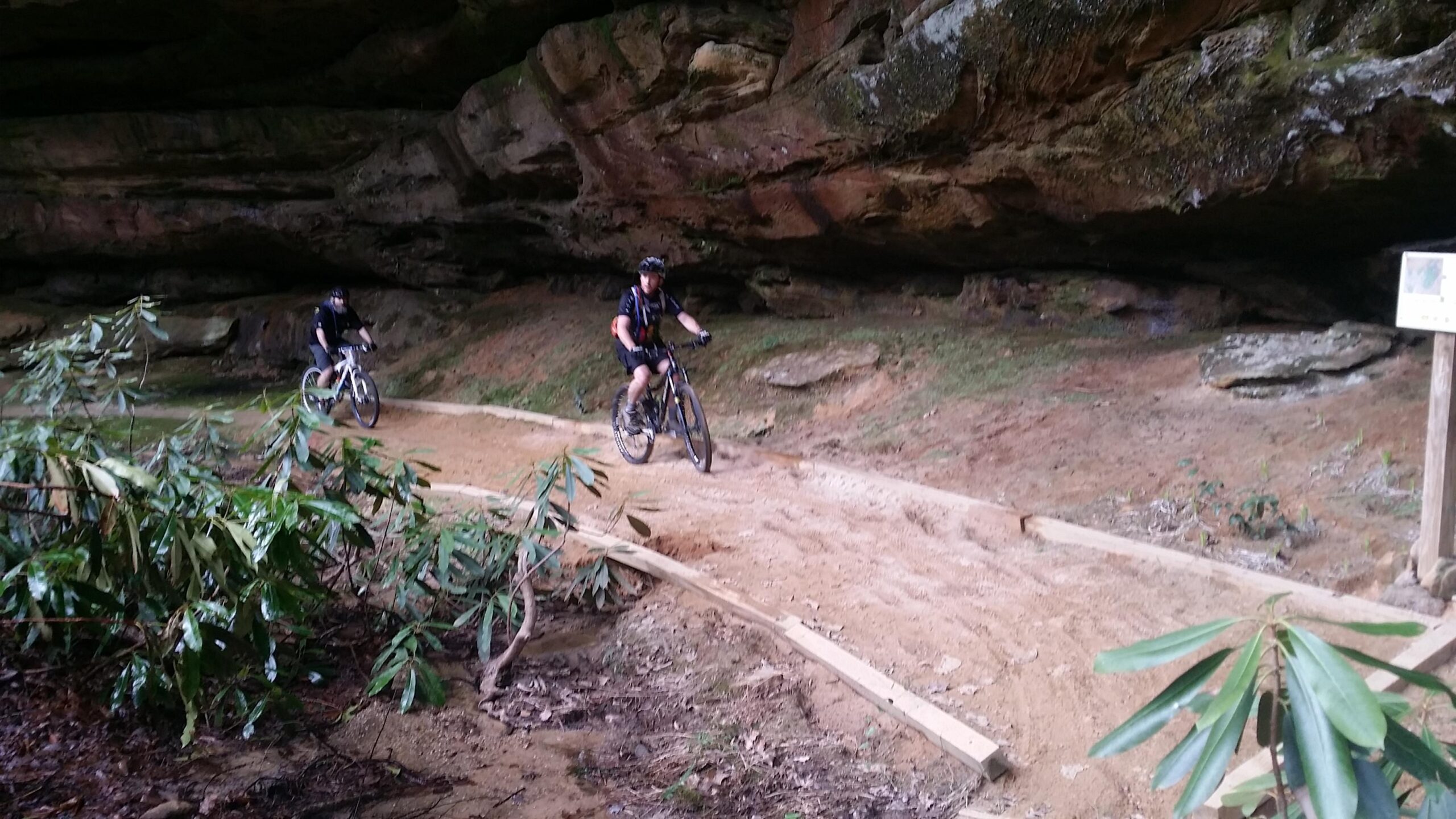 Two mountain bikers riding along a dirt trail beneath a rocky overhang, surrounded by lush green foliage and natural stone formations. The trail features wooden outlines and soft sandy areas, indicating a rugged outdoor environment. Big South Fork mountain bike trail.