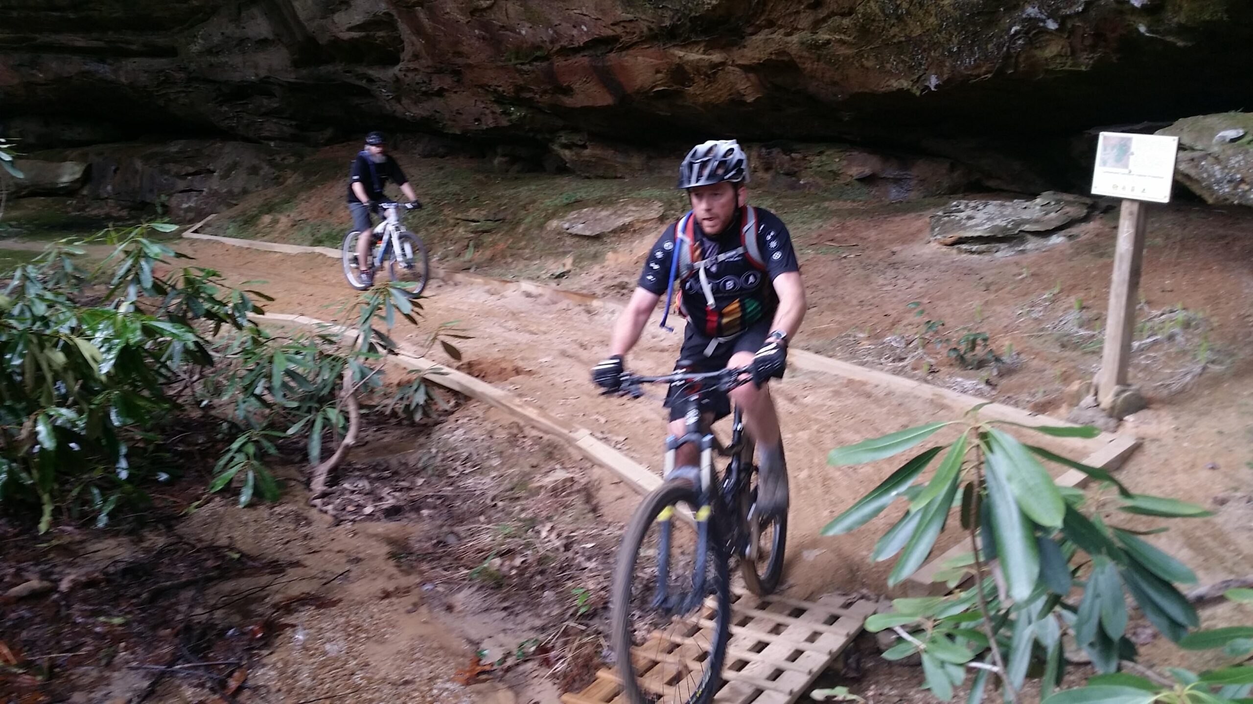 Two mountain bikers navigate a dirt trail under a rocky overhang, with one cyclist approaching a wooden bridge. Lush greenery and a sign are visible in the background. Big South Fork mountain bike trail.