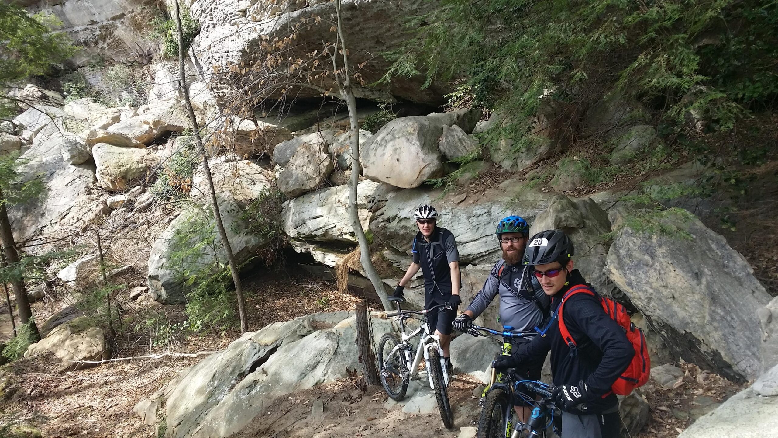 Three mountain bikers pose for a photo near rocky terrain and sparse trees. They are wearing helmets and biking gear, with two of them holding their bikes. The background features large rocks and a natural setting, suggesting an outdoor adventure. Big South Fork mountain bike trail.