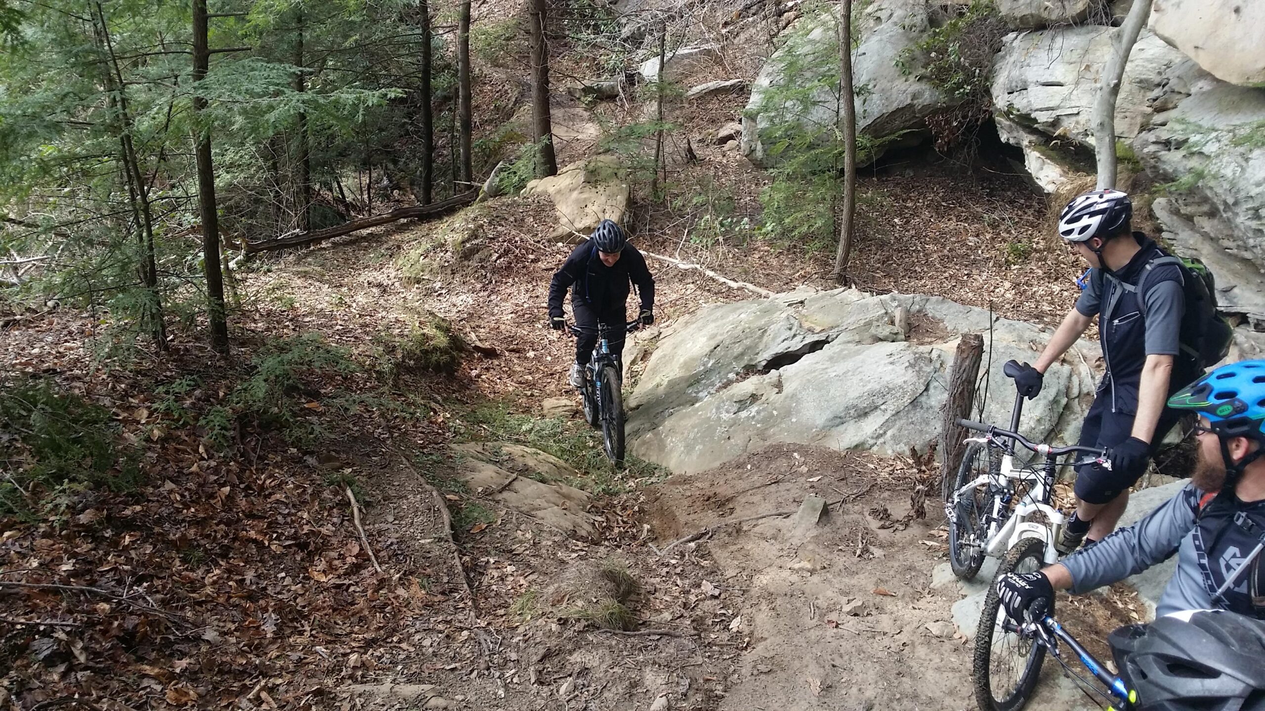 A mountain biker navigating a rocky trail surrounded by trees, with two other cyclists observing nearby. The scene depicts a forested environment with fallen leaves strewn across the ground and rugged terrain. Big South Fork mountain bike trail.