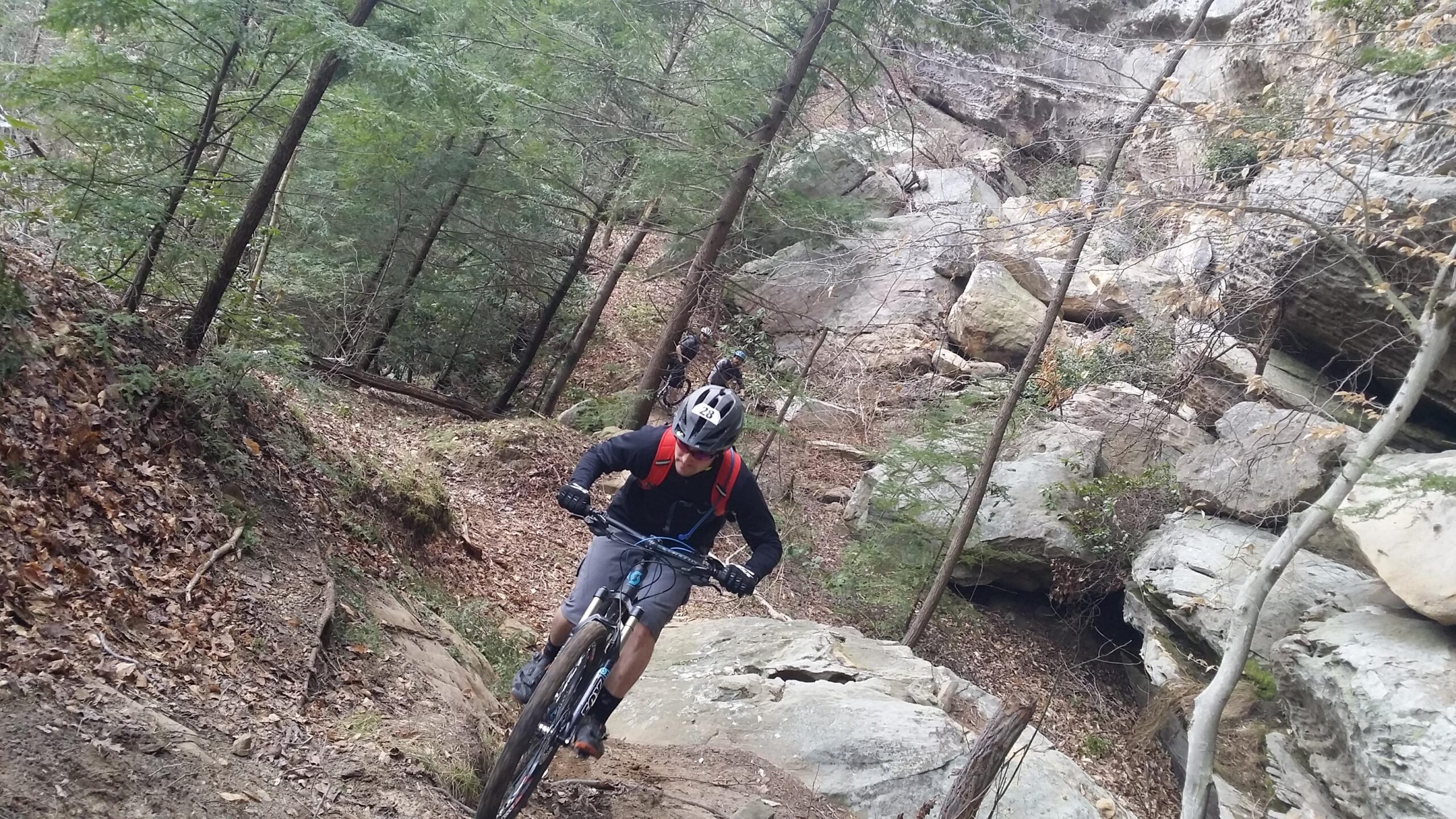 A mountain biker navigating a rocky trail in a forested area, surrounded by trees and large boulders. The rider is wearing a helmet and a backpack, focusing on the challenging terrain while another cyclist is visible in the background. Fallen leaves and dirt are present along the path. Big South Fork mountain bike trail.