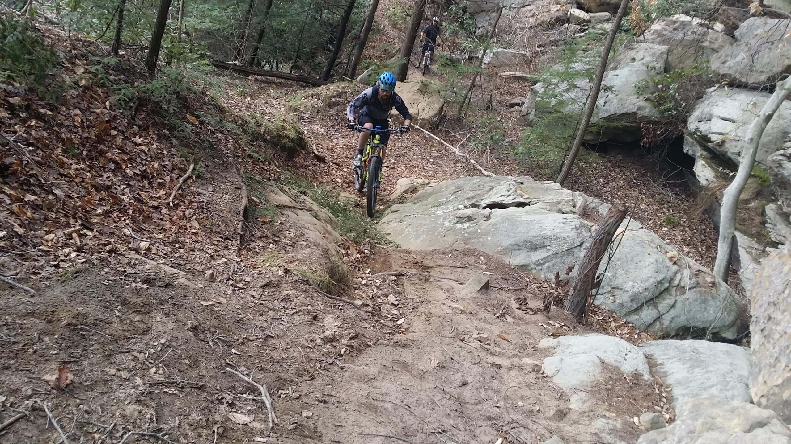 A mountain biker navigates a rocky and uneven trail surrounded by trees and fallen leaves. The path is steep with exposed rocks and dirt, showcasing a challenging terrain. In the background, another cyclist can be seen riding the trail. Big South Fork mountain bike trail.