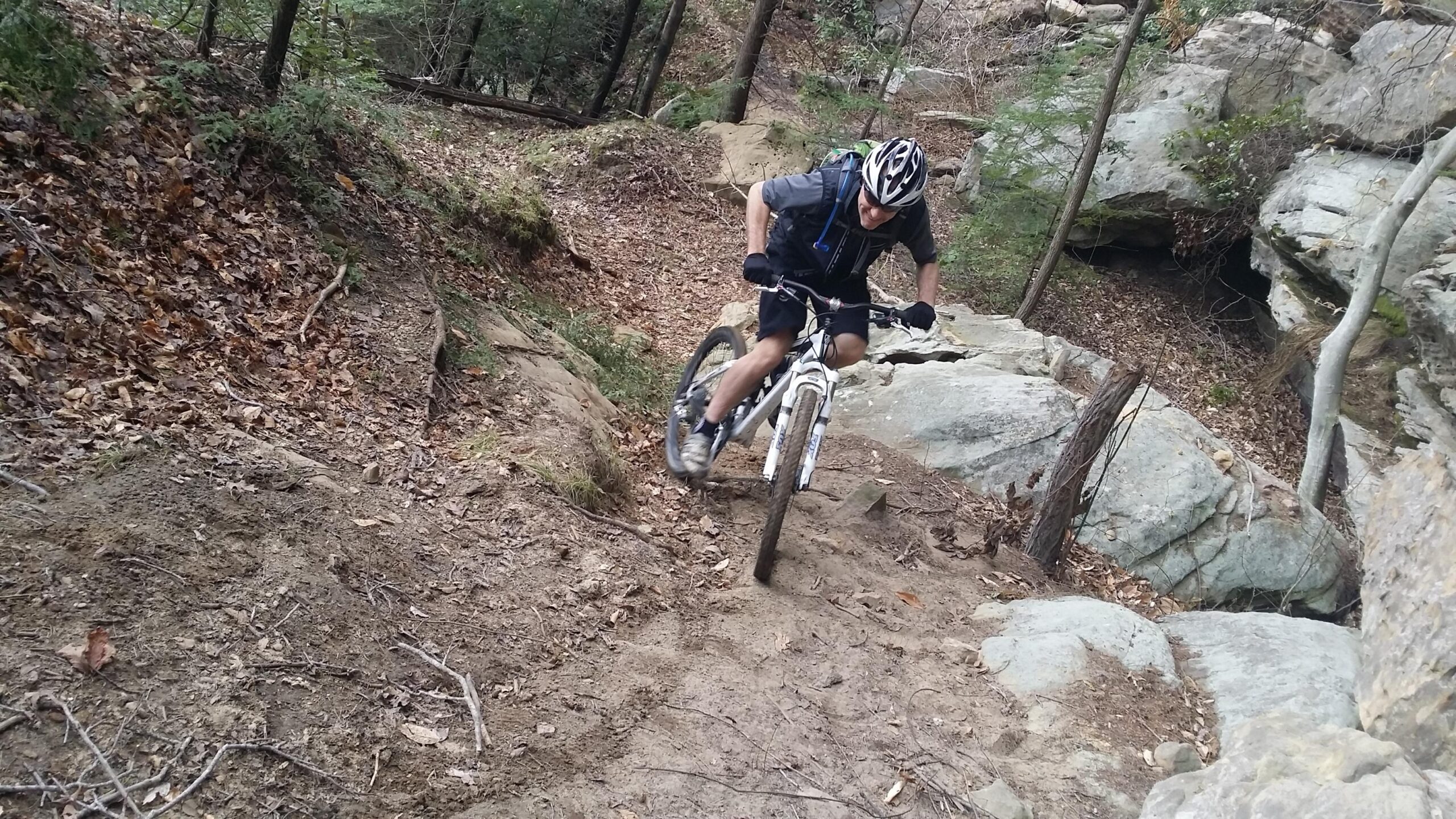 A cyclist maneuvering down a rocky trail surrounded by trees and autumn leaves. The rider is wearing a helmet and biking gear, focused on navigating the uneven terrain. Big South Fork mountain bike trail.