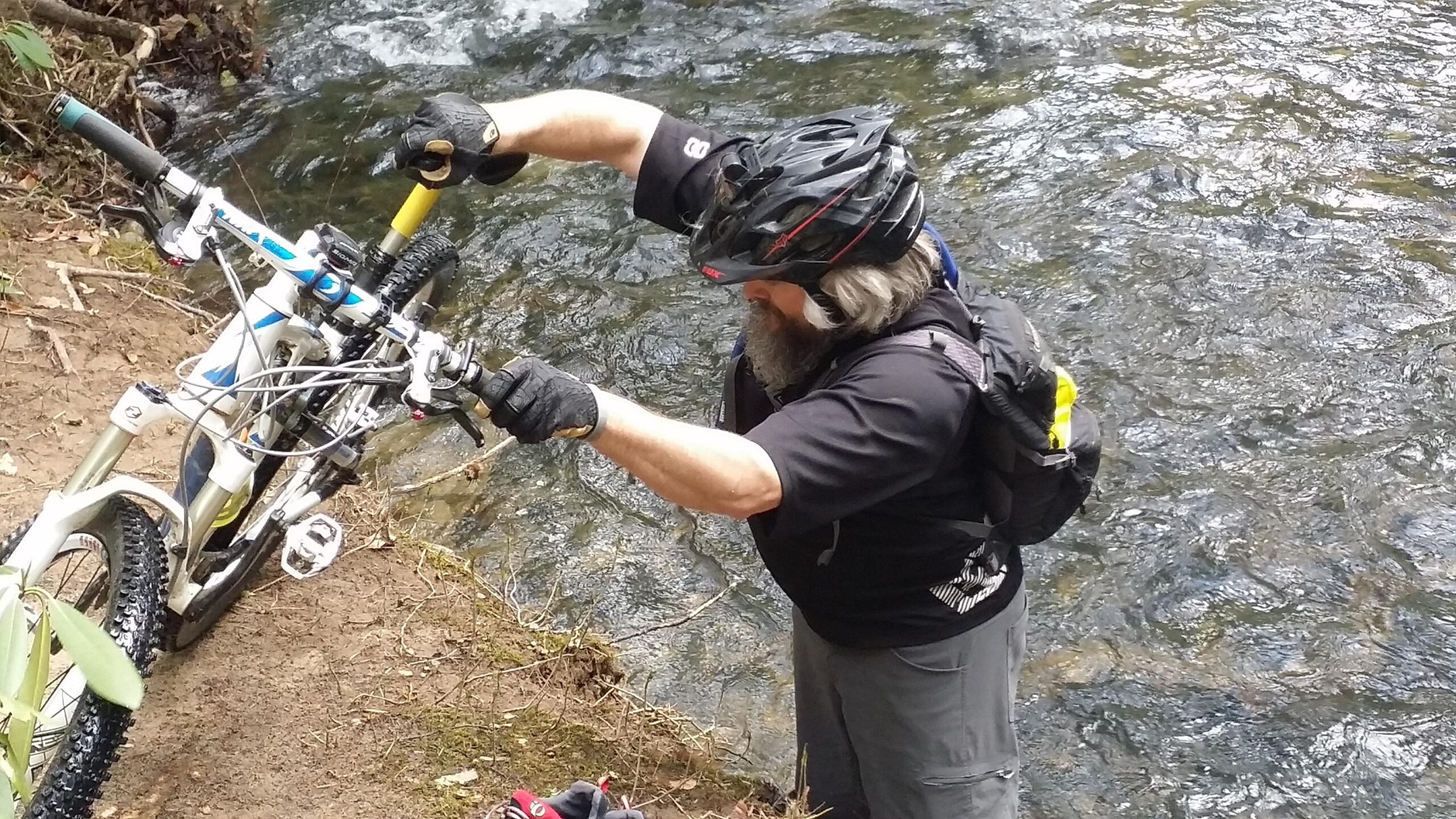 A person wearing a black helmet and gloves is fixing a mountain bike near a flowing creek. The bike is partially in the foreground, and the individual is focused on adjusting the handlebars while standing on the bank of the water. The surrounding area is lush with greenery. Big South Fork mountain bike trail.
