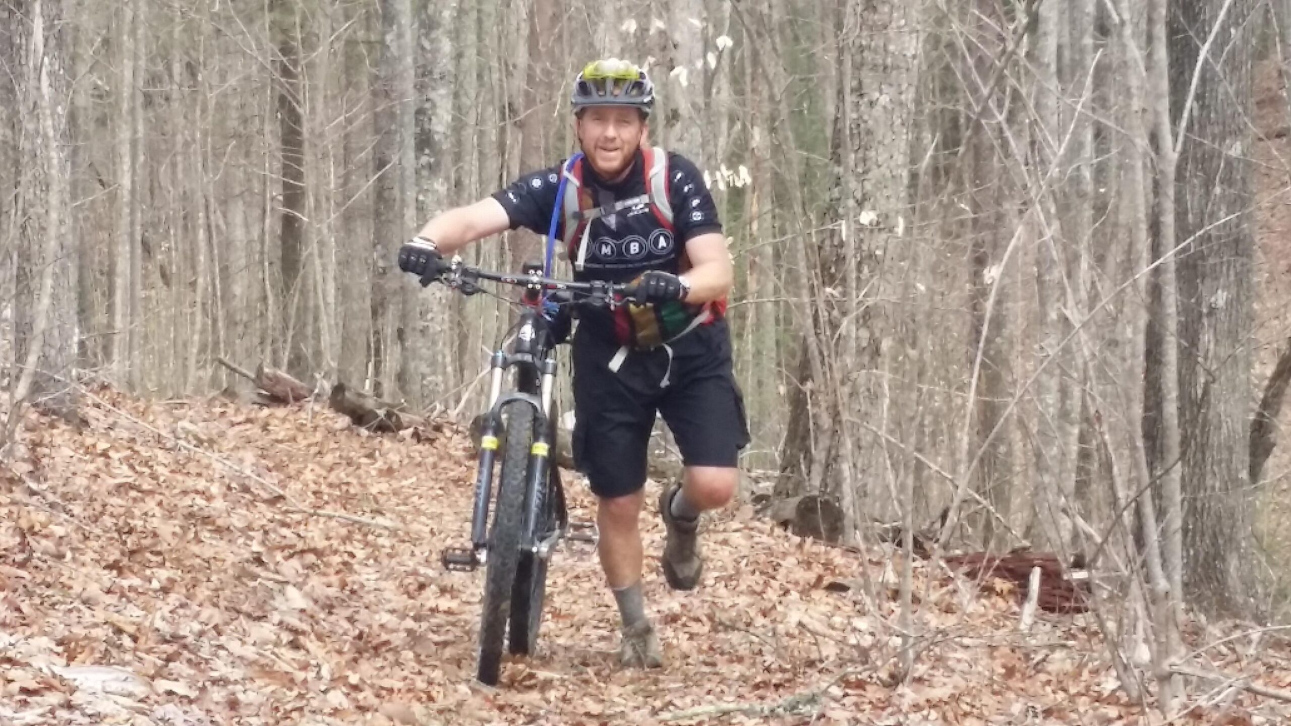 A mountain biker walking alongside his bike on a wooded trail covered in fallen leaves. He is wearing a helmet and athletic clothing, looking cheerful and engaged in the activity. Trees line the path, indicating a natural outdoor setting. Big South Fork mountain bike trail.