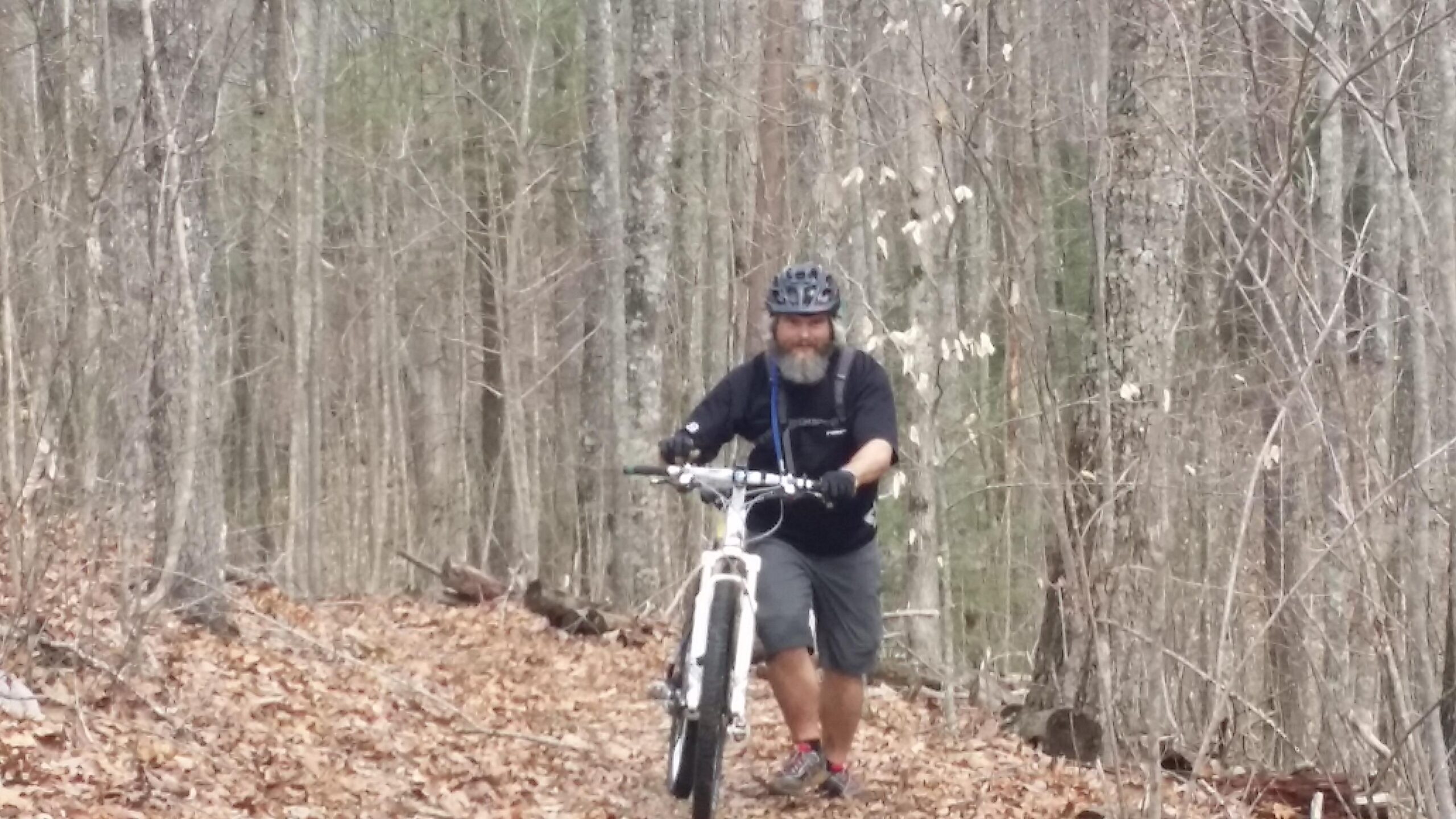 A person with a beard wearing a helmet and black shirt is pushing a mountain bike along a leaf-covered trail in a forested area with bare trees. Big South Fork mountain bike trail.