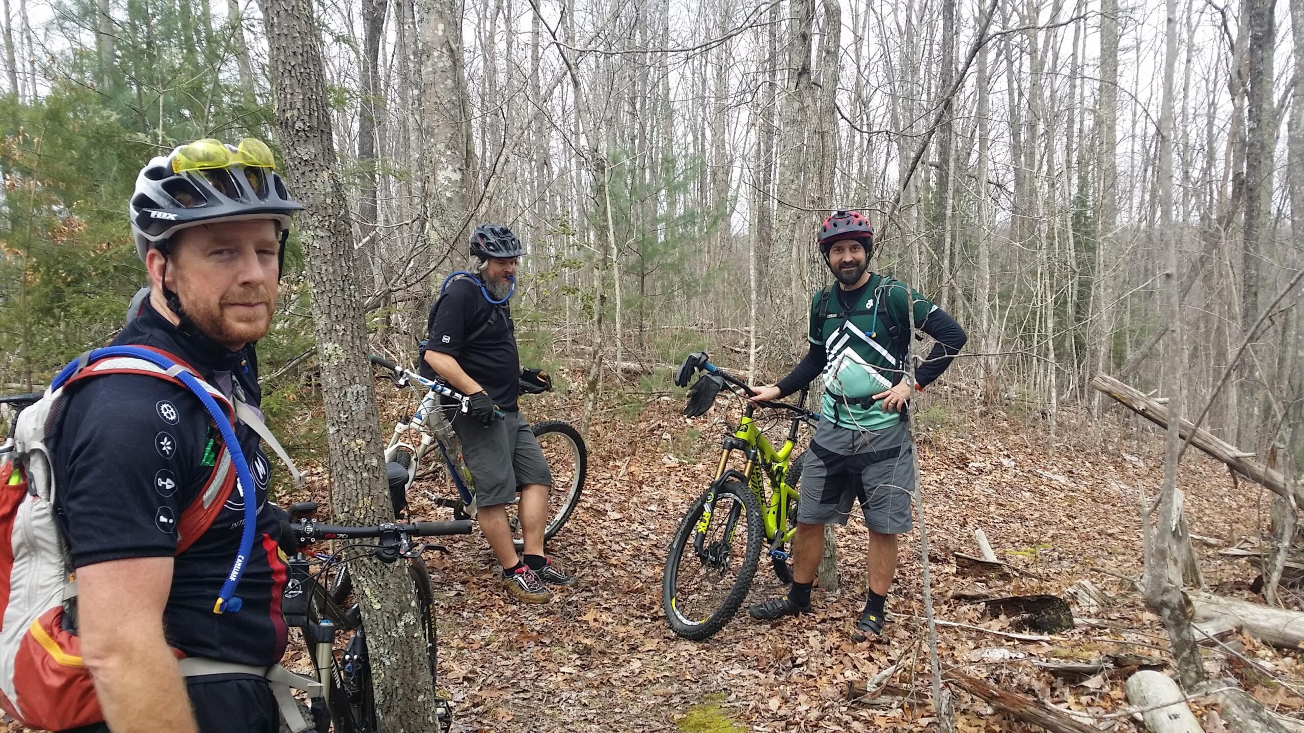 Three male mountain bikers pause on a dirt trail surrounded by trees and fallen leaves. They are wearing helmets and biking gear, with two standing beside their bikes while one leans against a tree. The atmosphere appears relaxed, with signs of a forested area in early spring. Big South Fork mountain bike trail.