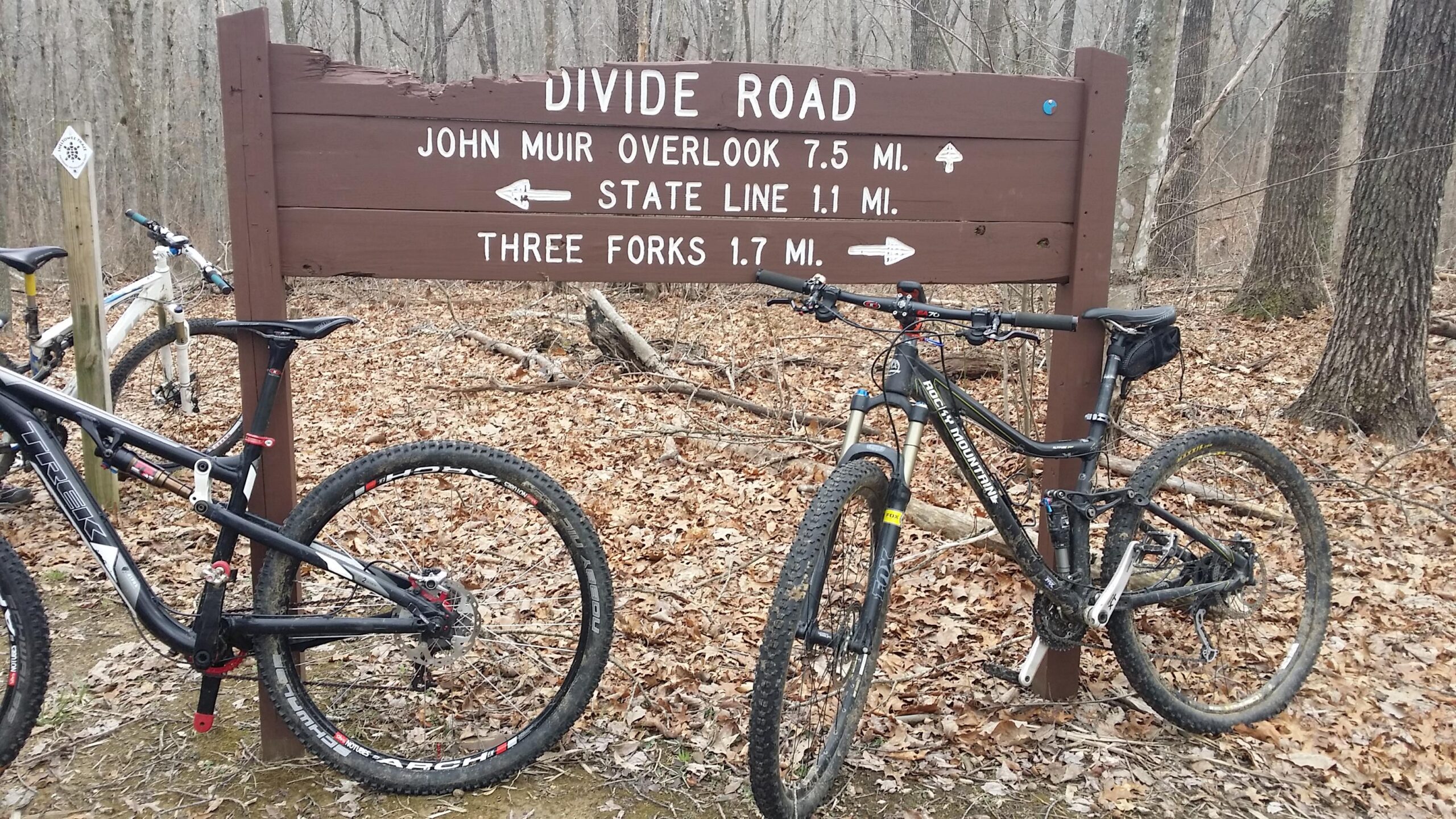 A signpost labeled "Divide Road" with directional arrows indicating distances to "John Muir Overlook" (7.5 miles), "State Line" (1.1 miles), and "Three Forks" (1.7 miles). Two mountain bikes are parked beside the sign, set against a backdrop of leaf-covered ground and bare trees. Big South Fork mountain bike trail.