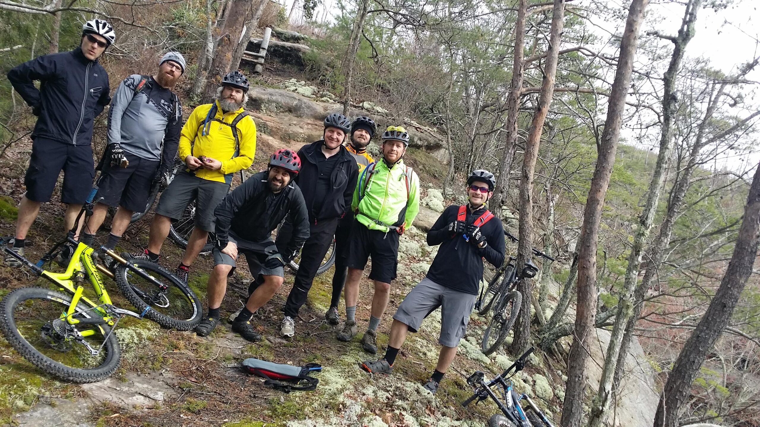 A group of eight mountain bikers poses for a photo on a rocky trail surrounded by trees. They are wearing helmets and cycling gear, and several bikes are leaning on the ground nearby. The scenery includes green moss and rocky terrain, indicating an outdoor adventure setting. Big South Fork mountain bike trail.