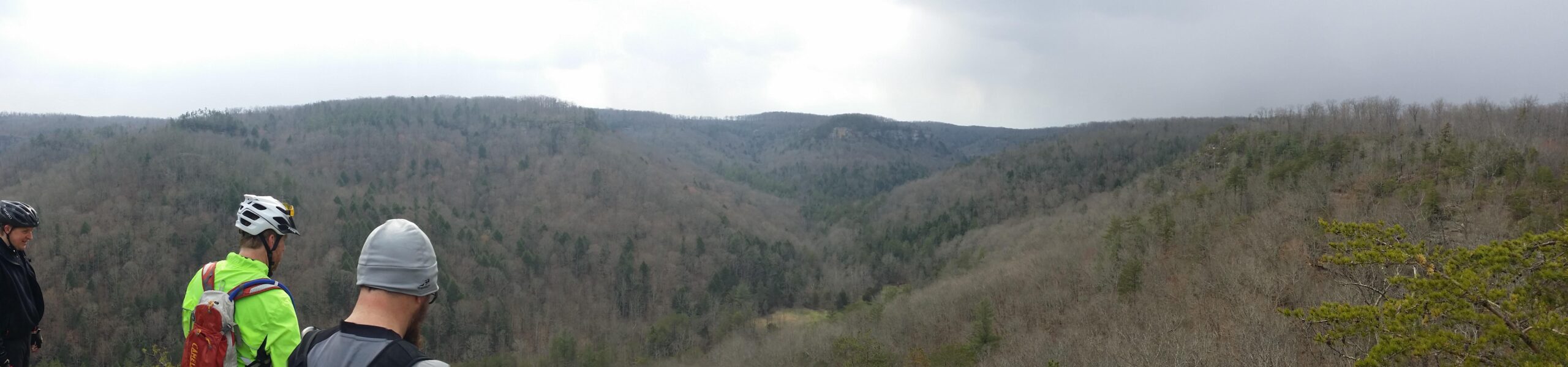 A panoramic view of a forested landscape with rolling hills under a cloudy sky. In the foreground, three people dressed in cycling gear are observing the scenery. The lush greenery of trees contrasts with the barren branches of other trees, indicating early spring or late winter. The scene conveys a sense of adventure and appreciation for nature. Big South Fork mountain bike trail.