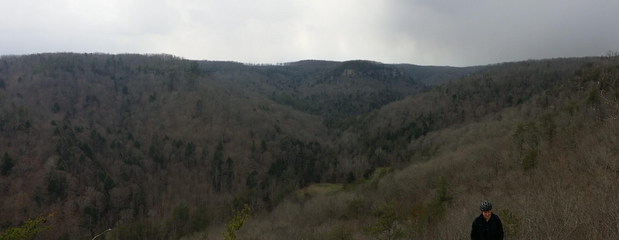 A panoramic view of a mountainous landscape during a cloudy day, featuring rolling hills with sparse trees in various shades of brown and green. In the foreground, a person wearing a helmet and a dark jacket stands on a rocky outcrop, looking down into the valley below. The distant hills extend into the horizon under a gray sky. Big South Fork mountain bike trail.