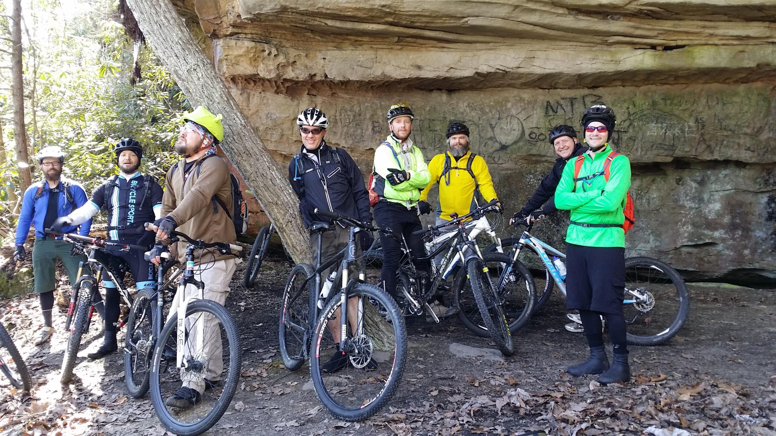 A group of eight mountain bikers gathered under a rocky overhang in a wooded area, some leaning against their bikes while others stand nearby. They are dressed in various cycling gear, including helmets and vibrant jackets, and appear to be enjoying a break in nature. The background features trees and a rocky wall with some graffiti. Fallen leaves are visible on the ground. Big South Fork mountain bike trail.