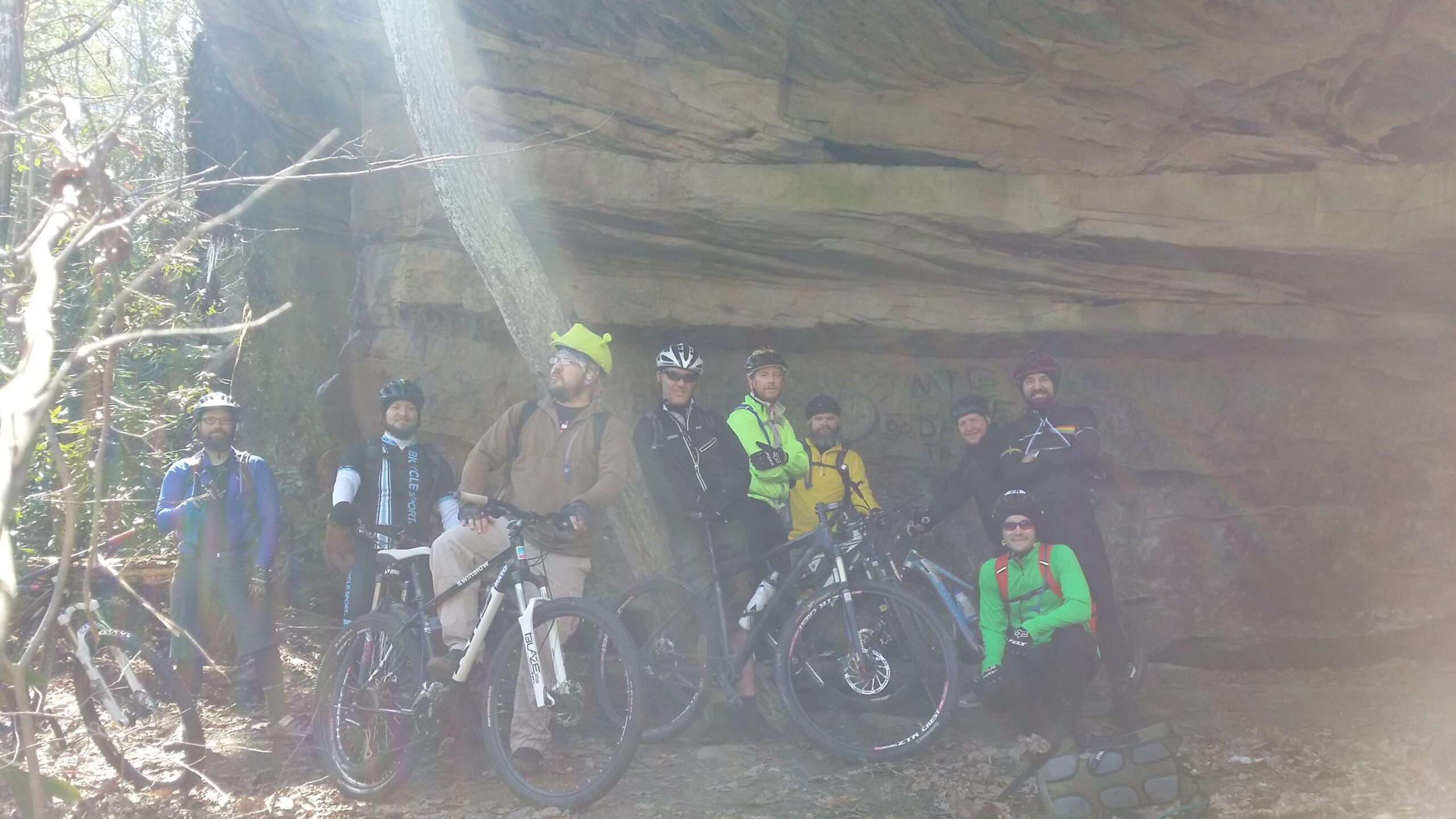 Alt text: A group of eight mountain bikers posing under a large rocky overhang in a wooded area. They are wearing various biking gear, including helmets and bright-colored jackets. Some bikes are leaned against the rocks, and there are trees and foliage in the background. Big South Fork mountain bike trail.