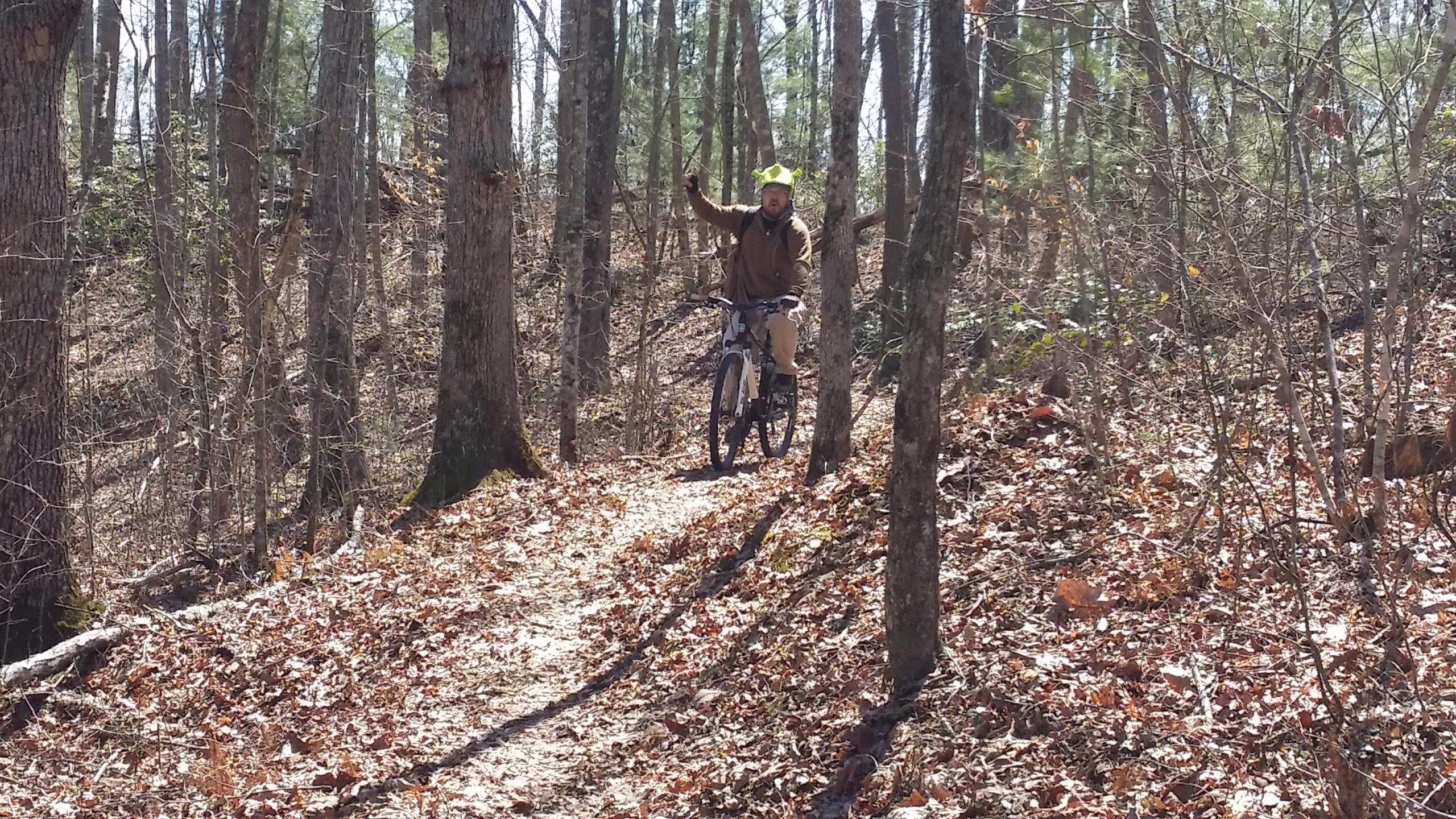 A person riding a mountain bike on a dirt trail surrounded by trees, with fallen leaves covering the ground. The rider is smiling and raising one hand in a gesture of excitement or greeting. The scene captures a sunny day in a wooded area. Big South Fork mountain bike trail.