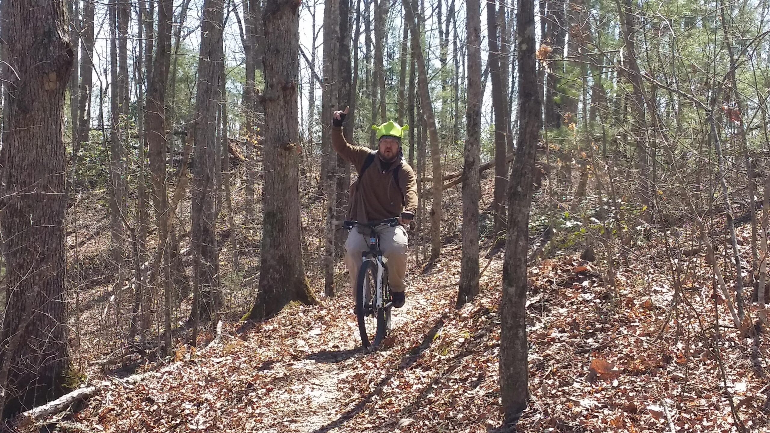 A person riding a mountain bike on a wooded trail, wearing a green hat resembling horns. The rider is raising one hand in excitement, surrounded by trees and fallen leaves, indicating a sunny day outdoors. Big South Fork mountain bike trail.