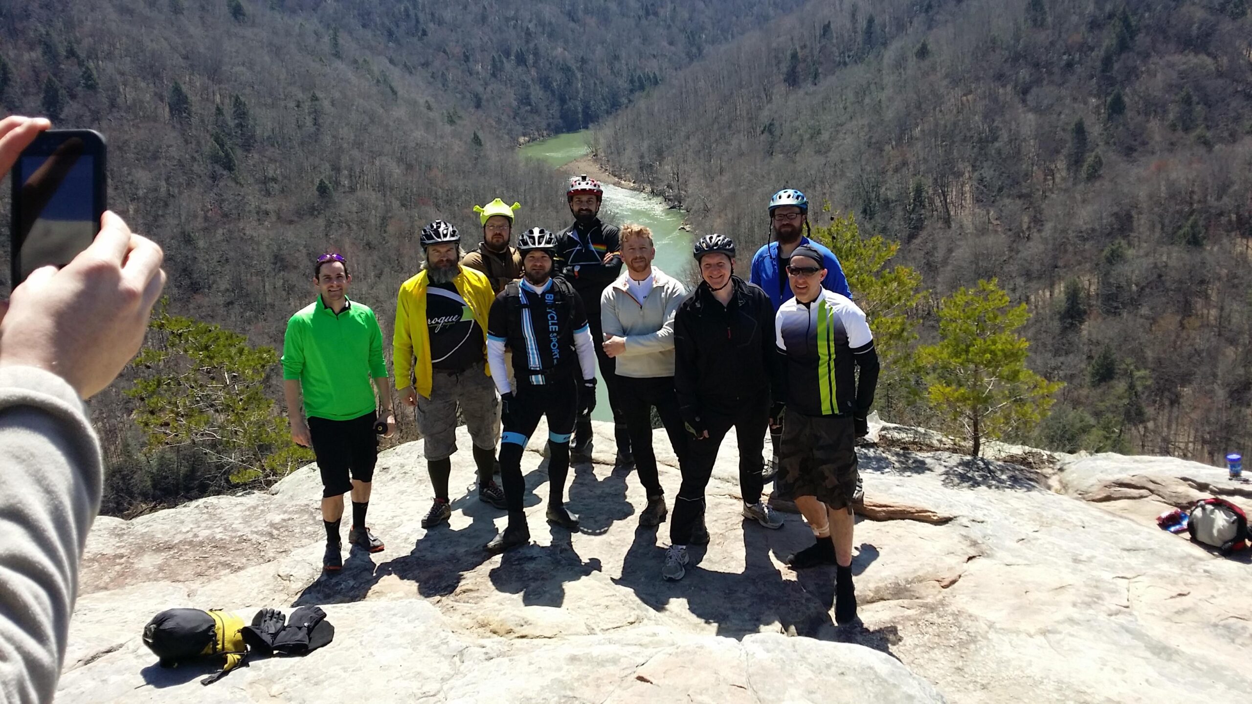 A group of eight individuals stands together on a rocky outcrop with a scenic view of a green river and forested hills in the background. They are dressed in outdoor attire, including cycling gear and casual clothing. One person is taking a photo of the group with a smartphone. The sky is clear and the weather appears sunny. Big South Fork mountain bike trail.