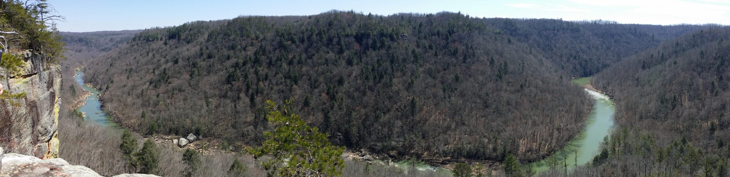 A panoramic view of a river winding through a wooded valley, surrounded by lush green hills and rocky cliffs. The landscape is mostly bare, suggesting early spring or late fall, with patches of evergreen trees dotting the hillsides. The water appears clear and reflects the sky above, creating a serene natural scene. Big South Fork mountain bike trail.