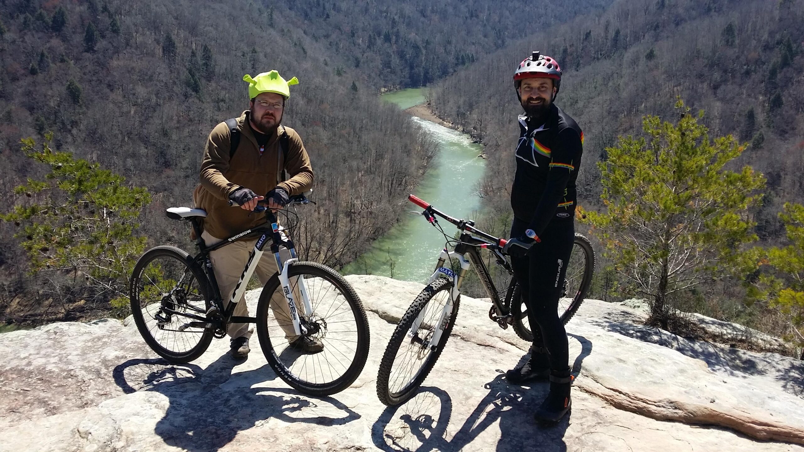Two mountain bikers stand on a rocky overlook, smiling at the camera. One rider, wearing a green helmet and a brown jacket, rests his hands on his bike. The other, dressed in a black cycling outfit with colorful accents, stands next to him. In the background, a winding river flows through a wooded valley, flanked by tree-covered hills under a clear blue sky. Big South Fork mountain bike trail.