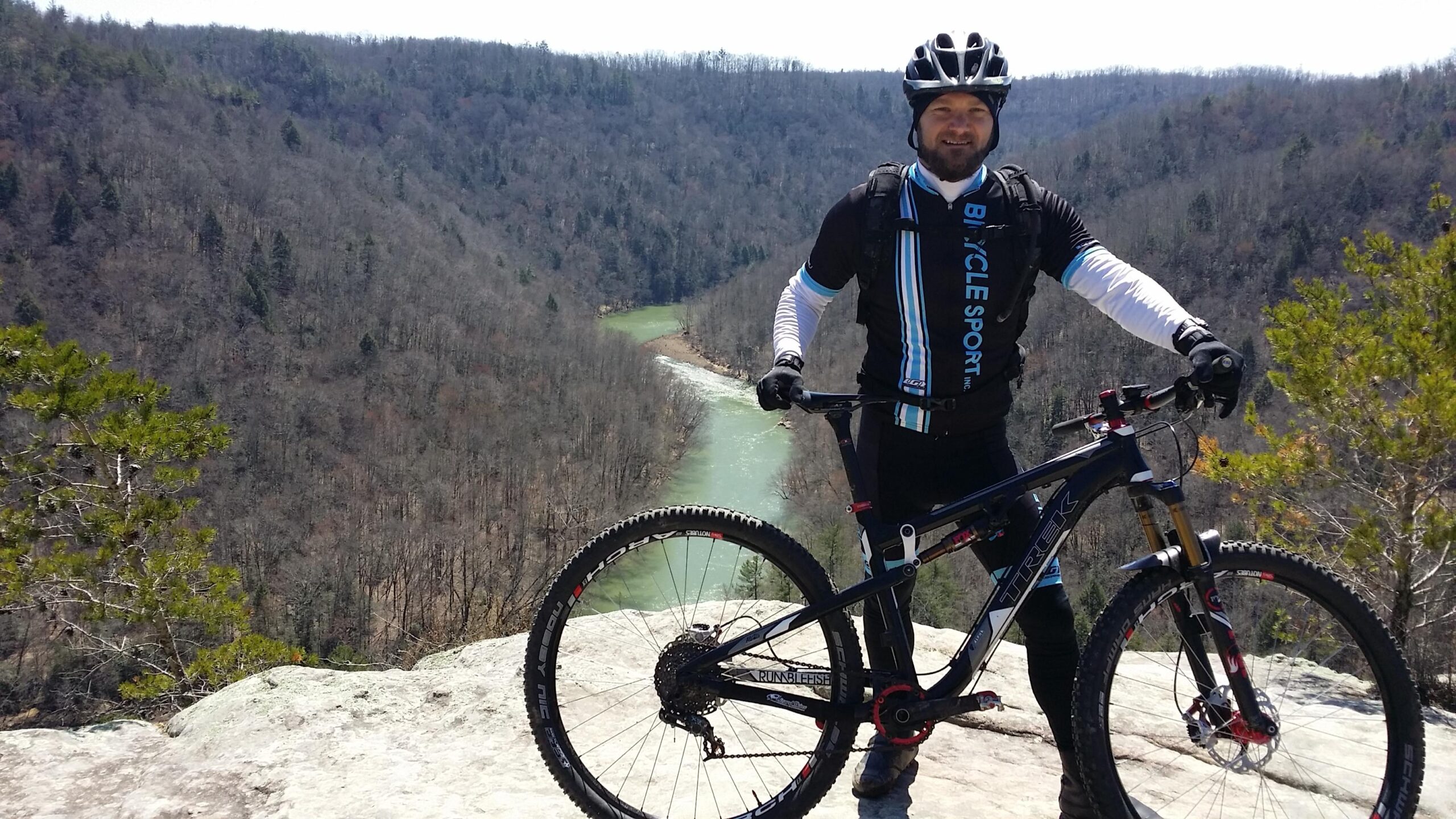 A mountain biker stands on a rocky outcrop overlooking a winding river surrounded by a forest of bare trees. The biker is wearing a black and blue cycling jersey, gloves, and a helmet, and is posing with a mountain bike. In the background, the landscape features rolling hills and a clear blue sky. Big South Fork mountain bike trail.