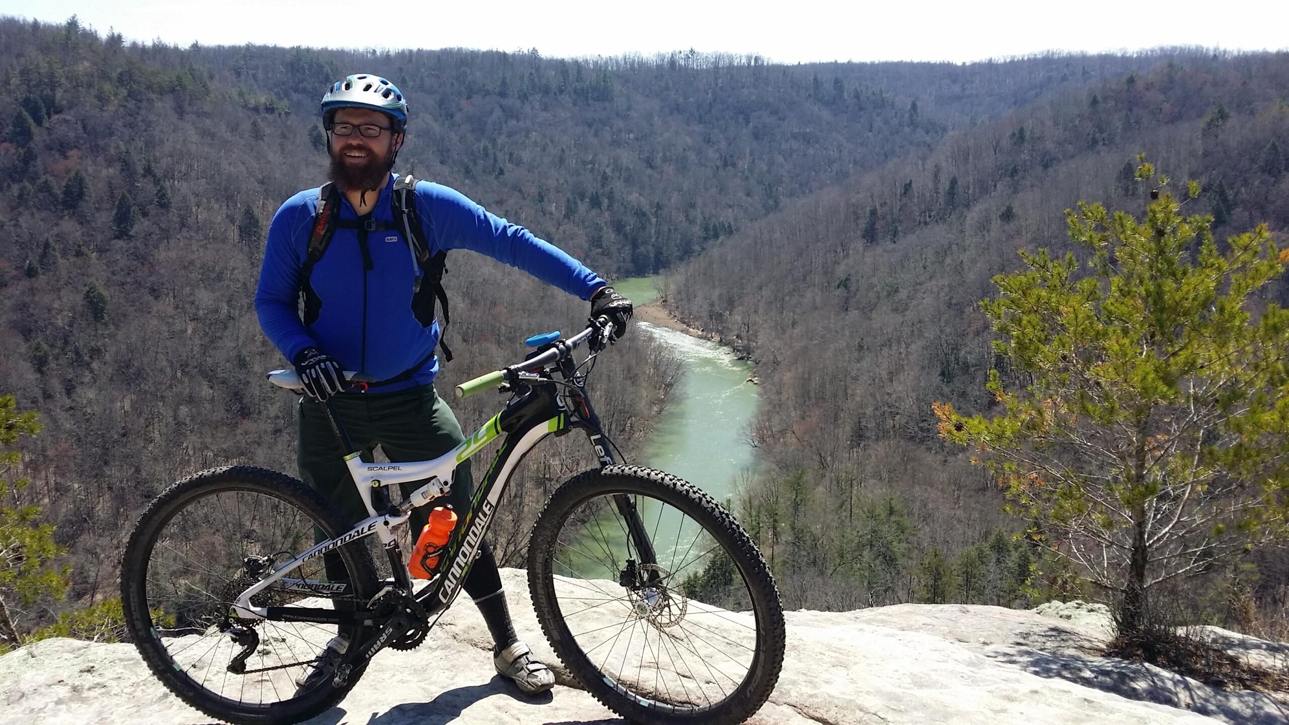 A person in a blue long-sleeve shirt and biking gear stands on a rocky overlook, smiling while holding a mountain bike. In the background, a flowing river winds through a valley surrounded by trees, showcasing a scenic landscape under a clear sky. Big South Fork mountain bike trail.