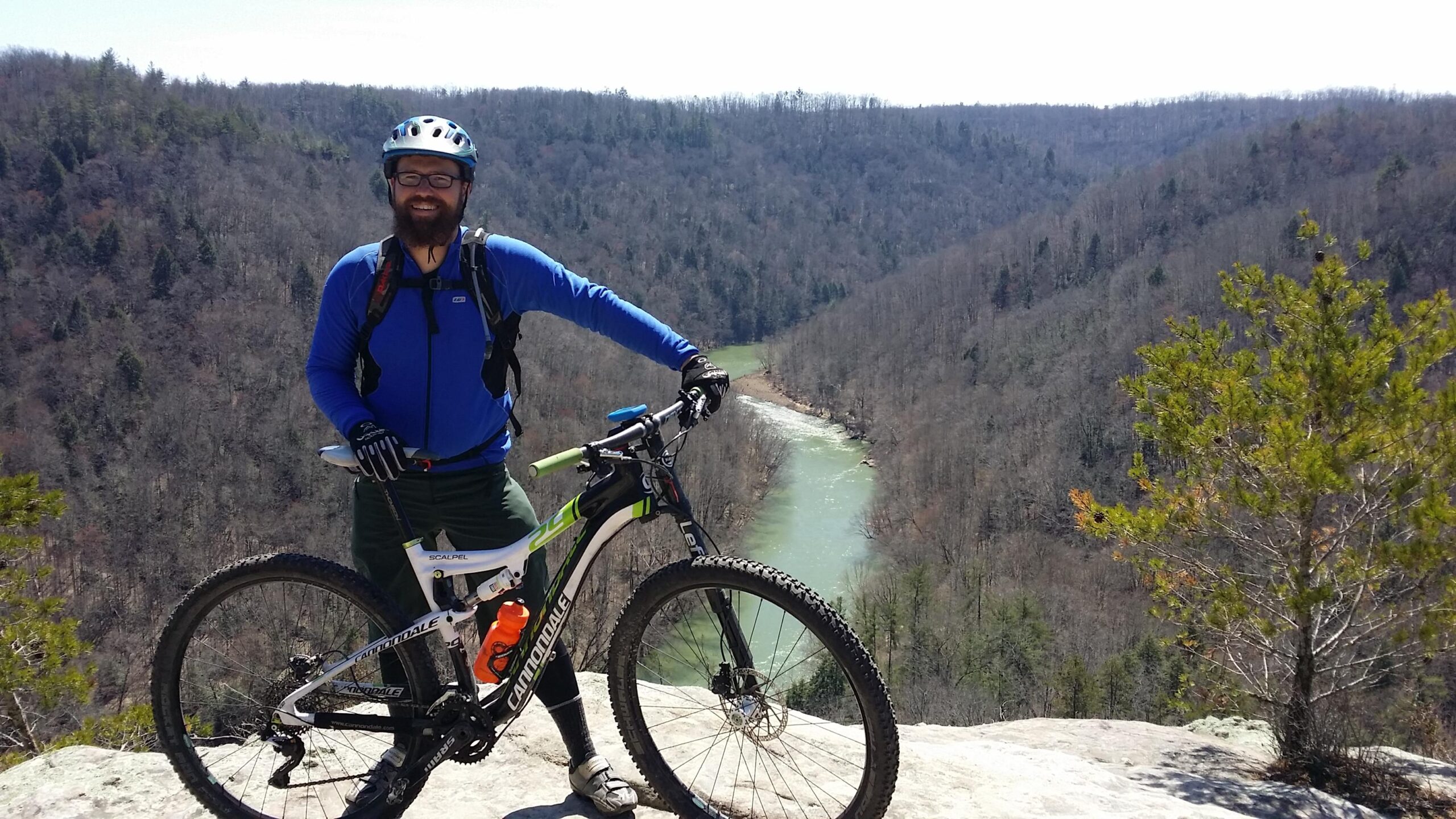 A man wearing a blue long-sleeve shirt, helmet, and gloves stands next to a mountain bike on a rocky overlook. The background features a river winding through a valley surrounded by trees in a mountainous landscape. The scene is bright and sunny, reflecting a day of outdoor adventure. Big South Fork mountain bike trail.