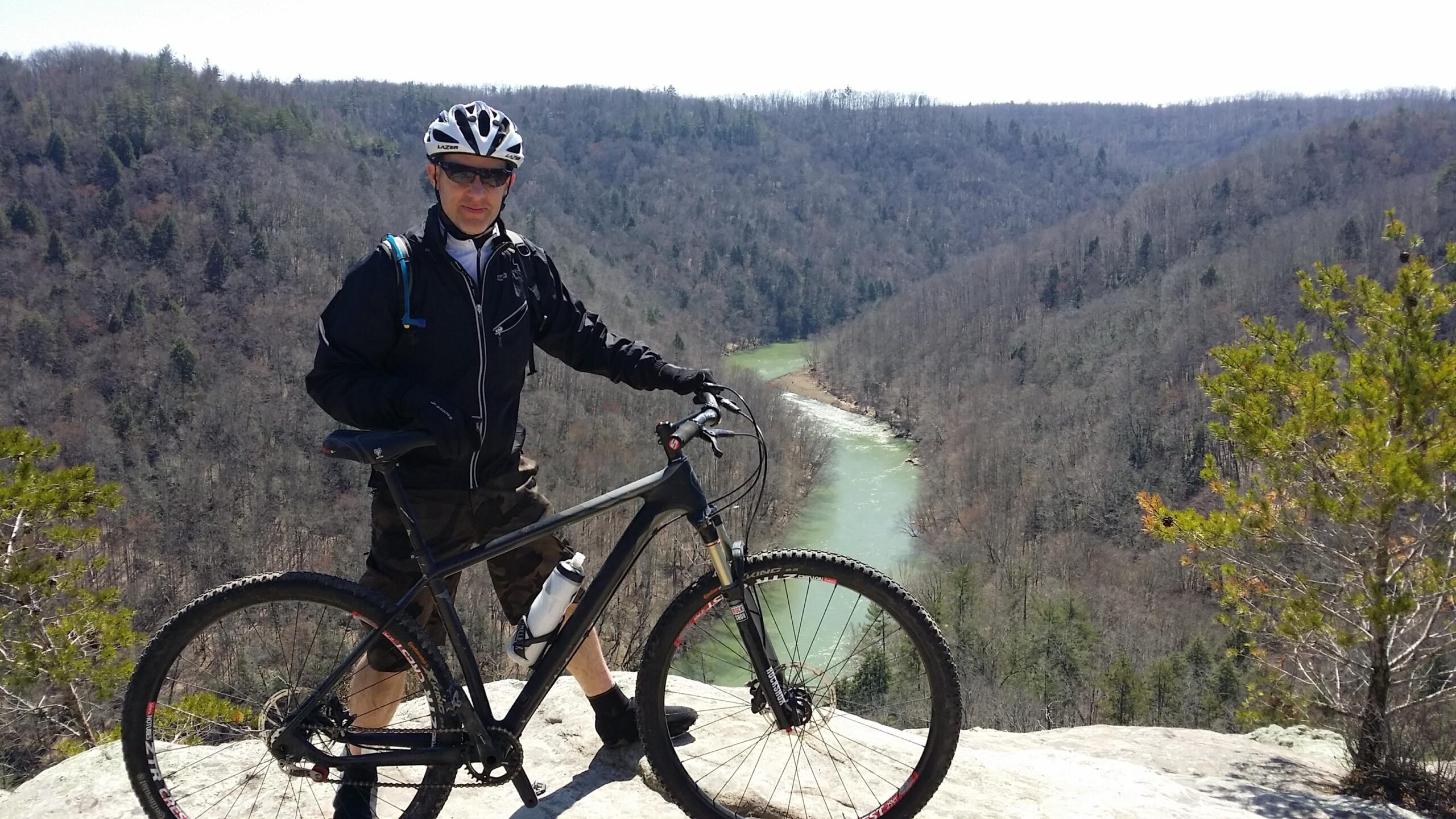 A mountain biker stands on a rocky outcrop overlooking a winding river surrounded by trees. The cyclist is wearing a helmet and sunglasses, dressed in a black jacket and camouflage shorts, while his bike rests beside him. The landscape features rolling hills and a clear blue sky. Big South Fork mountain bike trail.
