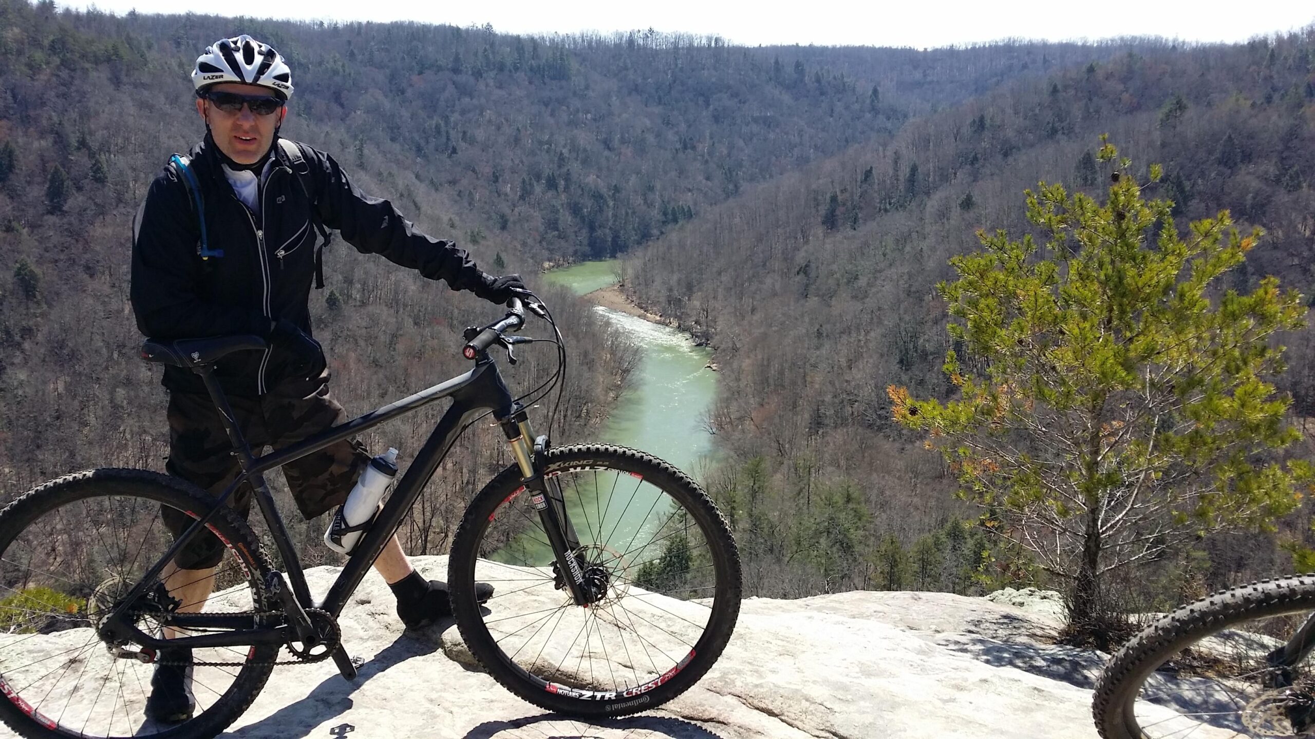 A person in outdoor gear, including a helmet and sunglasses, stands next to a mountain bike on a rocky viewpoint overlooking a winding river surrounded by a forested landscape. The scene captures early spring colors, with trees still bare of leaves in the background. Big South Fork mountain bike trail.